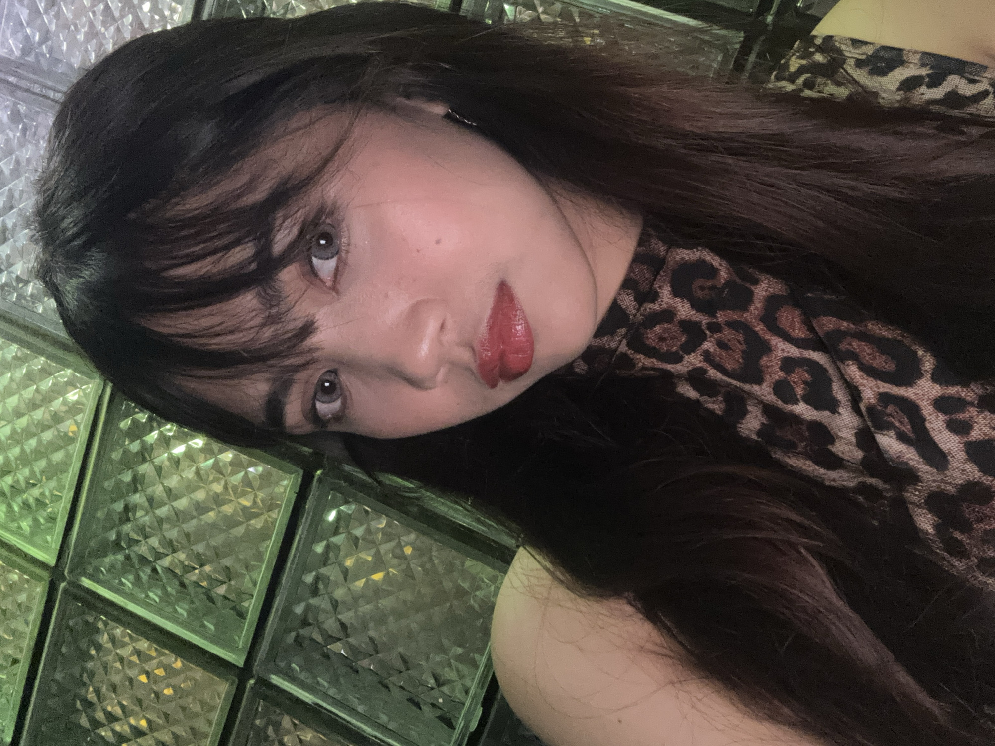 The image is a selfie of a young woman with long, dark hair and bangs. She is wearing a leopard print top. She has red lipstick on and appears to be looking at the camera. The background is a wall of textured glass blocks, lit with a green glow. There is no specific indication of the location within Davao City, Philippines.