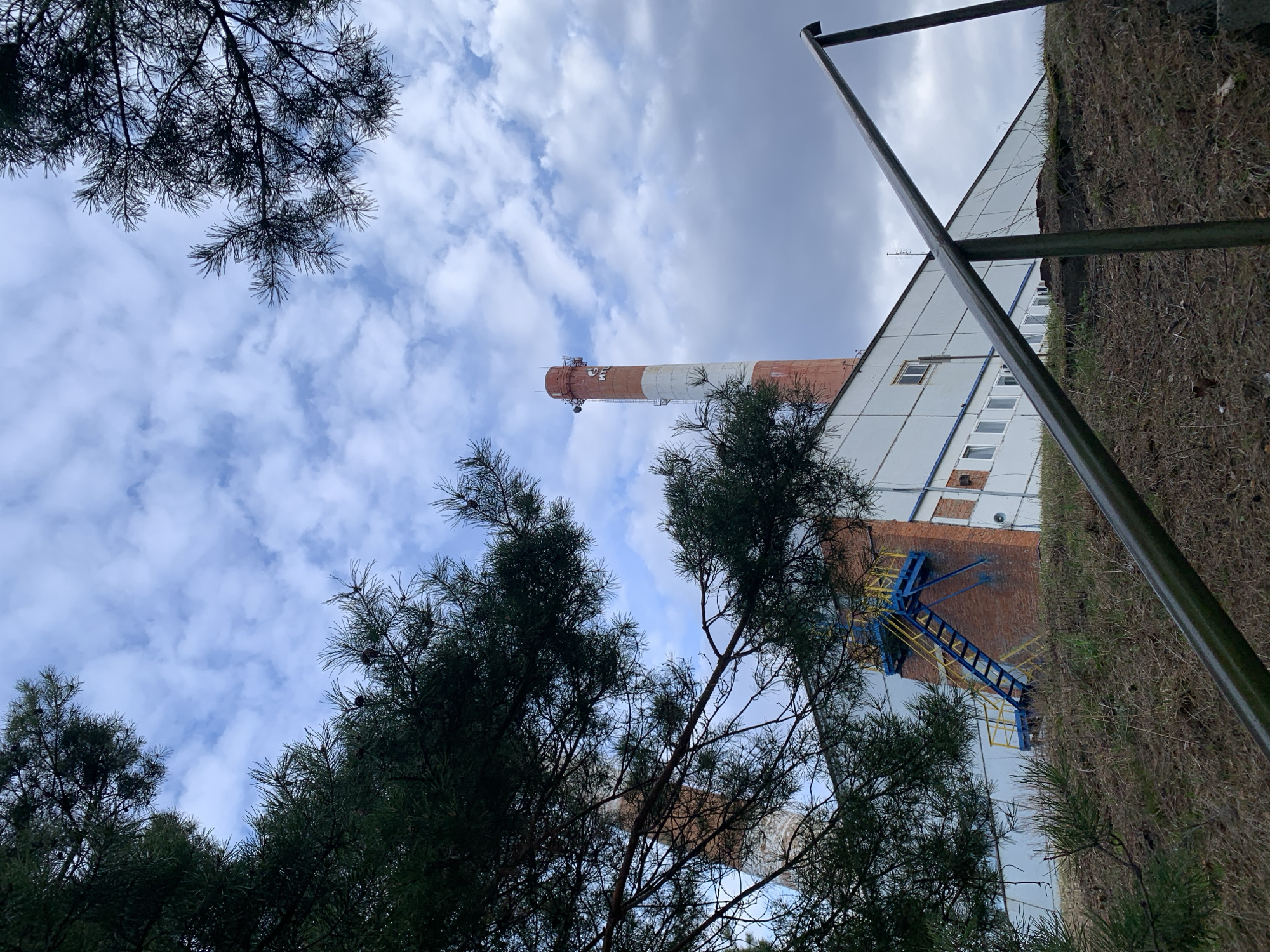 The image, taken from a low angle, depicts a building and a smokestack in Chaikovsky, Russia. The building is a mix of brick and white siding, with a blue and yellow ladder visible on the brick section. The smokestack is painted with alternating red and white bands. The sky is cloudy, with some patches of blue visible. Trees, particularly pine trees, are in the foreground, partially obscuring the view. An angled metal pole is in the upper right corner of the image. The ground to the right of the building appears to be an embankment covered in dry vegetation. No people are visible in the image.