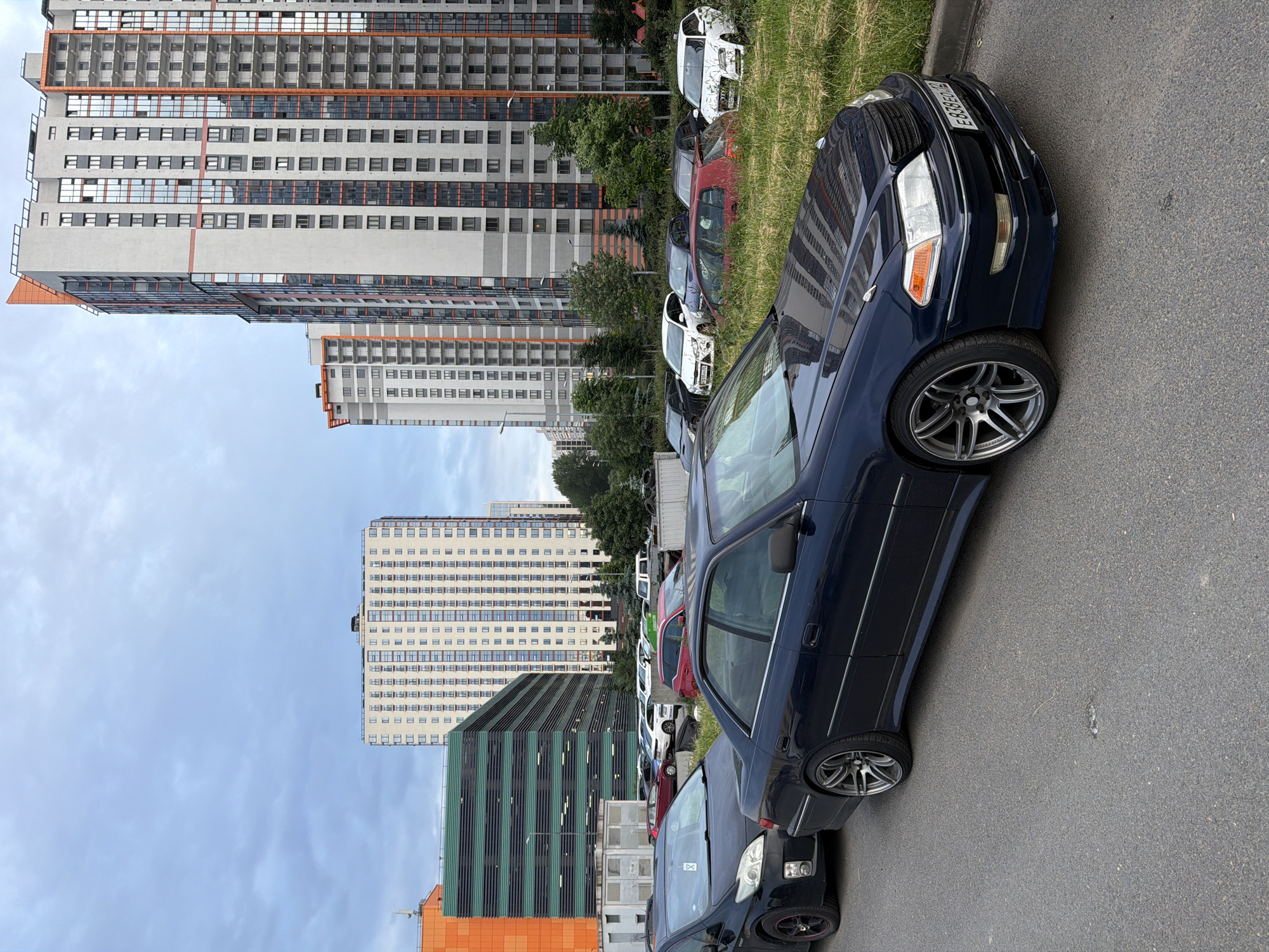In an urban environment, a dark blue sedan is parked on a paved road. The car is positioned in the foreground, angled towards the right of the frame, with its front passenger-side wheel visible. Behind the sedan, several other cars are parked on a grassy area. The background features modern, multi-story residential buildings with a mix of white, grey, and orange facades, as well as a multi-level parking structure with a green and orange exterior. The sky is overcast.