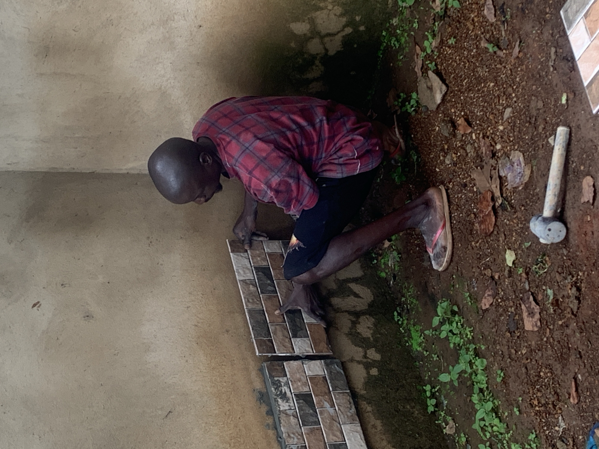 A bald Black man is kneeling on the ground and tiling a wall in Agban, Nigeria. He is wearing a red and black plaid shirt and dark pants, with flip-flops on his feet. He is placing a small, rectangular tile on a wall that is partially tiled with similar mosaic tiles. The wall appears to be made of rough concrete and is stained with dirt and possibly mildew. The ground in front of the wall is covered with dark brown soil, scattered leaves, and small green plants. A hammer lies on the ground to the right of the man. The scene is outdoors, with natural light.