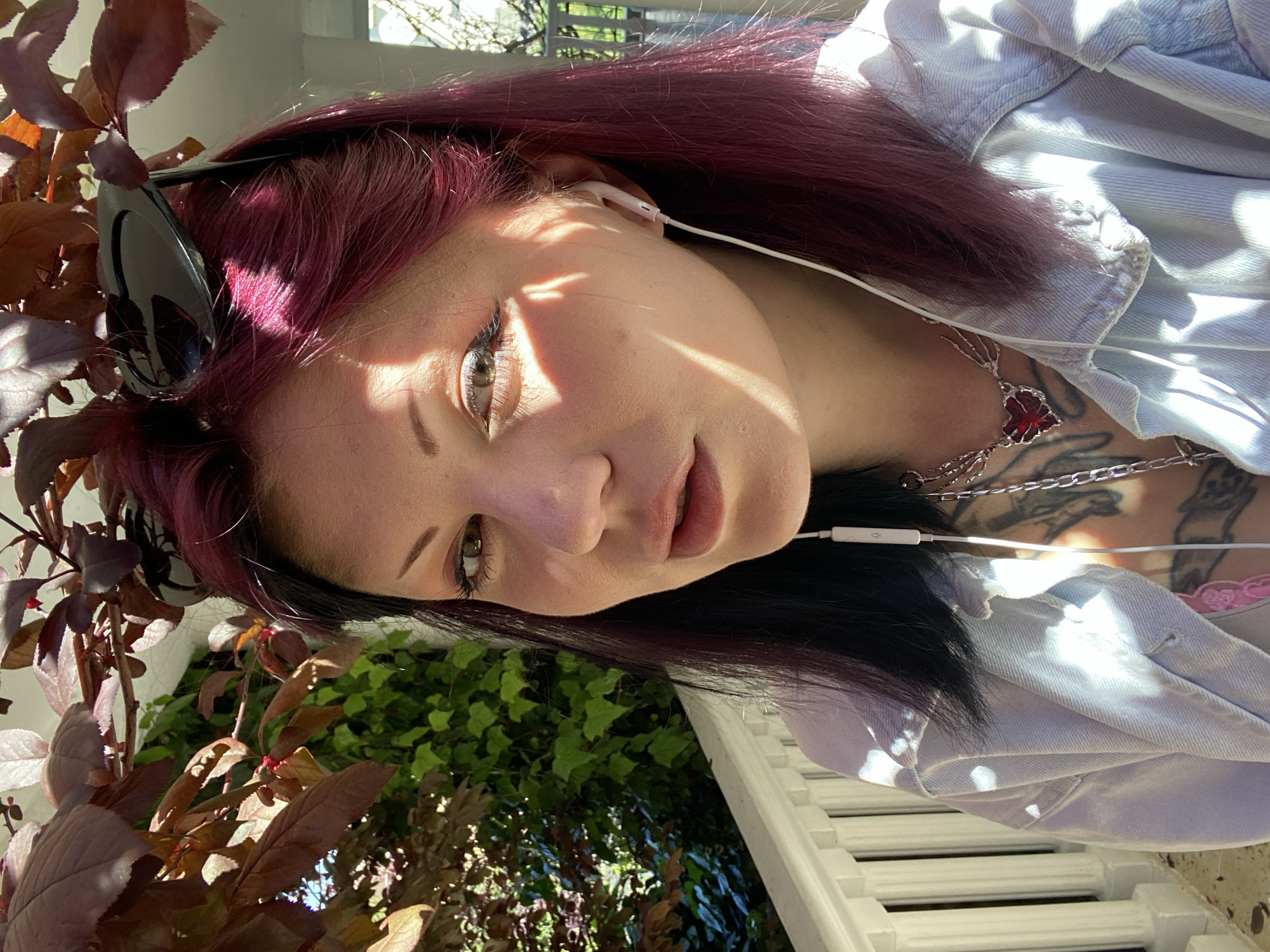 A young woman with dyed burgundy hair is the focal point of the image. Her hair transitions to black at the roots. She is wearing white earphones and has sunglasses perched atop her head. She also has necklaces and visible tattoos on her chest. Her eyes are hazel. She is wearing a light blue denim jacket. She is situated outdoors. In the foreground, there are reddish-brown leaves of a tree or shrub. In the background, there is green foliage and a white railing, which suggests the location may be a balcony or patio. The image is taken in bright sunlight, casting strong shadows across her face. The location of Kunje, Montenegro, cannot be verified from the image.