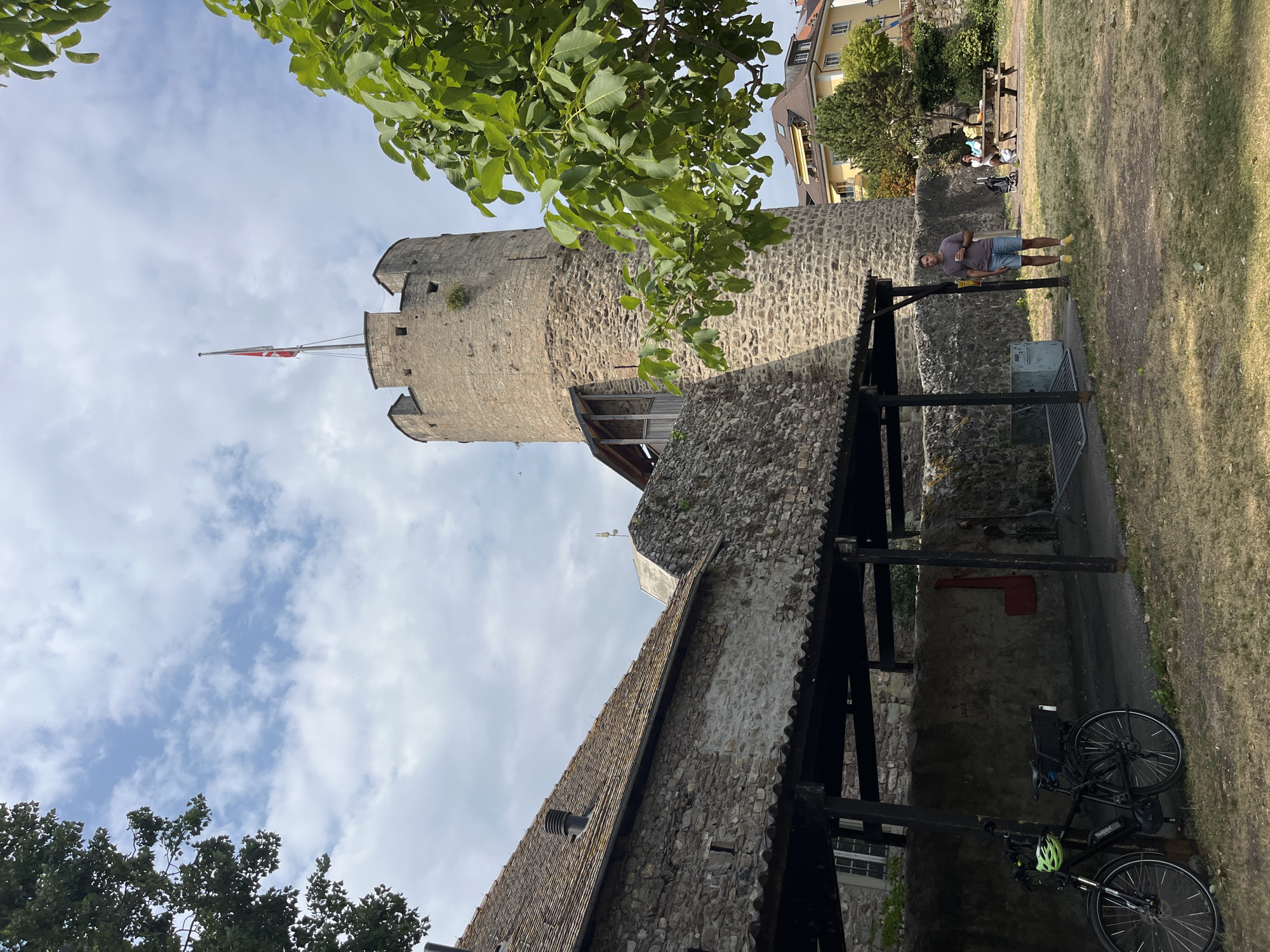In the city of La Tour-de-Peilz, Switzerland, a stone tower stands tall against a partly cloudy sky. The tower features a flag at its summit. To the left of the tower, a stone building with a tiled roof is visible, featuring an overhang supported by wooden posts. In the foreground, a black bicycle with a green helmet is parked under the overhang. A man wearing a t-shirt and shorts stands in the middle ground, holding a drink. In the background, a picnic table is occupied by two people, and a yellow building with a brown roof is situated behind a tree. The ground is covered with dry grass.