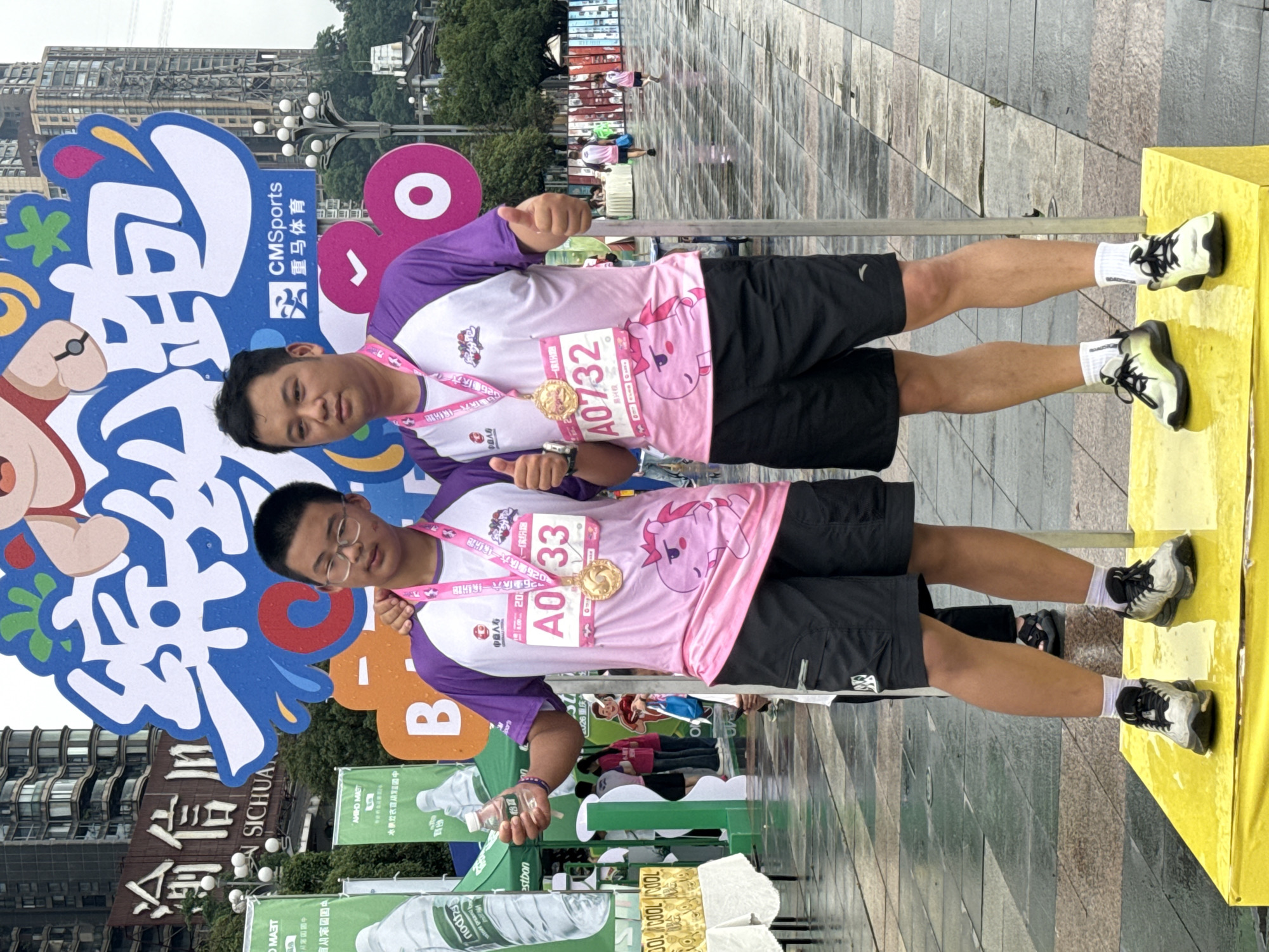 Two men are posing for a photo after an event; they are standing in front of signage from "CMSports 亚马体育," wearing purple and pink shirts with numbers "A0732" and "A0/33" pinned to them and gold medals around their necks. They are giving a thumbs up. The ground is paved, and in the background is a partially visible building with a sign saying "SICHUA." To the left are banners and items that appear to be water bottles and paper towels, and the edge of a yellow object. The city is Nan'an District, China.