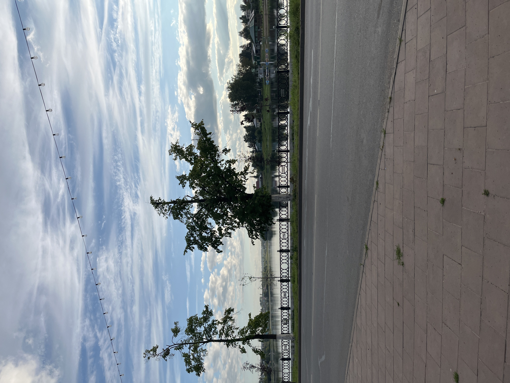 The image shows a waterfront scene in Blagoveshchensk, Russia, likely along the Amur River embankment. In the foreground, a paved sidewalk made of reddish-brown rectangular bricks runs diagonally from the bottom left to the middle of the frame. Adjacent to the sidewalk is a paved road with a white lane marking. Beyond the road, a decorative black metal fence with an intricate circular pattern separates the embankment from a body of water, which is the Amur River. Several trees are visible along the embankment, including two prominent ones with full green canopies. Across the river, the opposite bank is lined with buildings, trees, and possibly some boats or structures on the water. The sky is blue with wispy white clouds, and a string of decorative lights is suspended across the upper portion of the frame. The overall impression is a peaceful, scenic urban waterfront.
