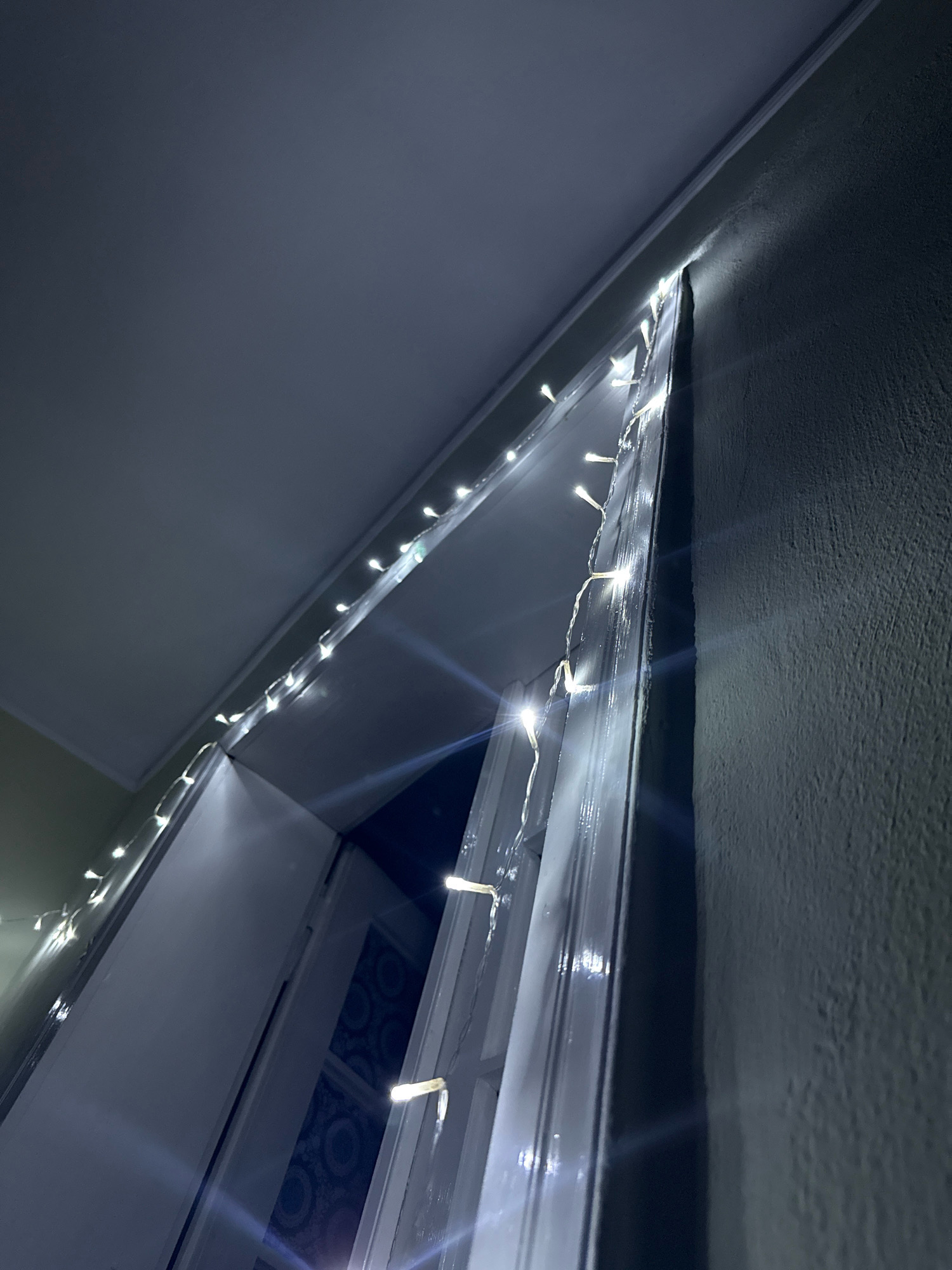 In an indoor environment, a string of white fairy lights is draped along the top edge of a white doorframe. The lights are small and illuminate the frame with a soft glow. The doorframe itself appears to be a glossy white, reflecting the light. To the right of the doorframe, a textured, dark gray wall is visible, showing slight imperfections in its surface. Above the doorframe, a smooth, dark gray ceiling slopes downwards, meeting the wall at a sharp angle. The overall scene is dimly lit, with the fairy lights serving as the primary source of illumination.
