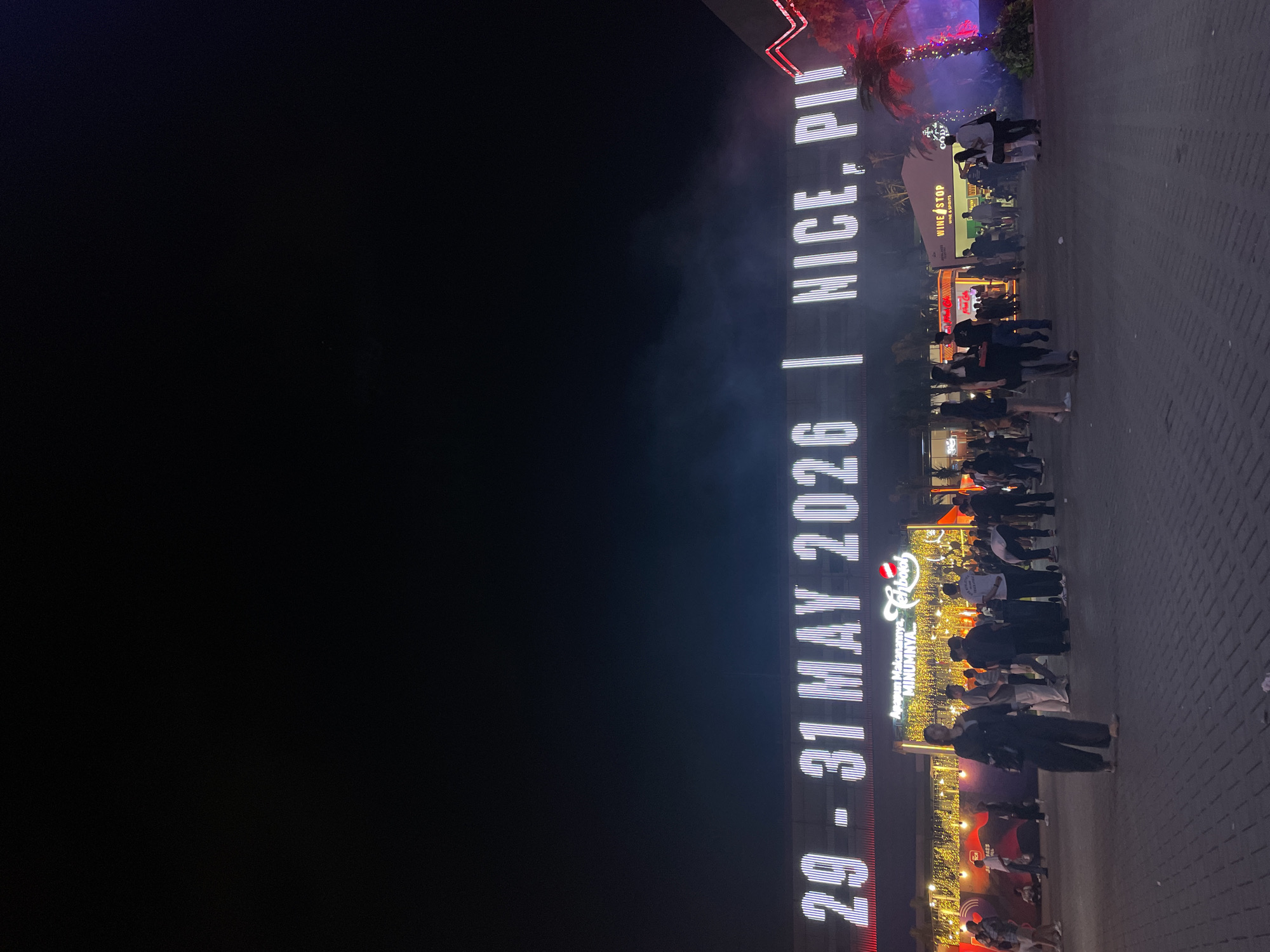 The image shows a night scene in Salembaran, Indonesia, where a crowd of people gathers near an illuminated vertical sign reading "29-31 MAY 2026 | NICE, PI." The sign also features an advertisement for a restaurant or food-related establishment with the text "Apapun Makanannya, MINUMNYA" and the name "Tenbord." A "WINE STOP WINE & SPIRITS" storefront is visible behind the crowd. Palm trees adorned with lights add to the nighttime ambiance. The foreground shows paving stones.