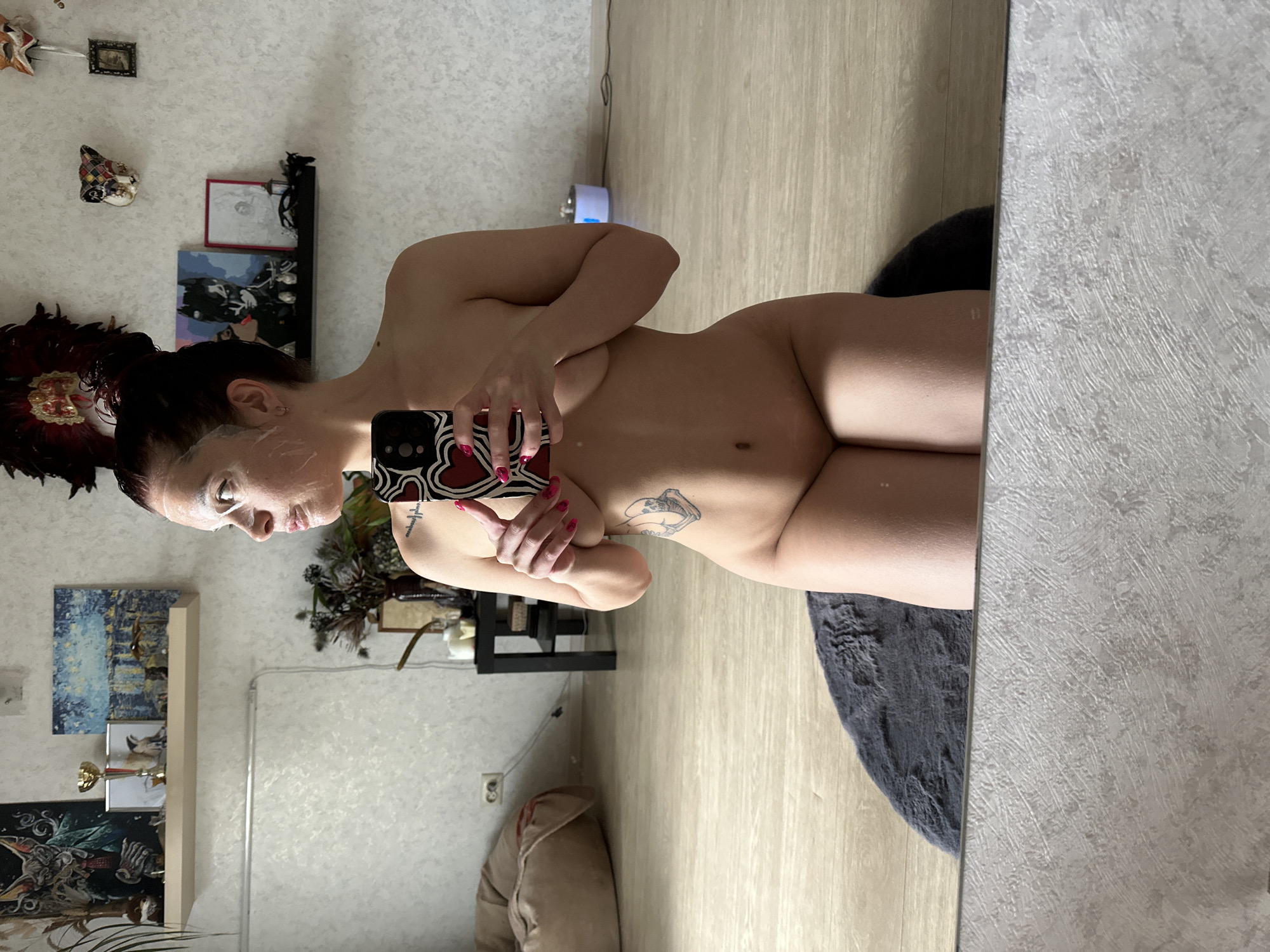 A nude woman is positioned in front of a mirror, capturing a selfie with her smartphone. She is partially covered by her arms, holding the phone in her right hand, and her left arm is visible. She has tattoos on her arm and torso. Her face is covered in a white substance, possibly a mask. Behind her, there is a wall decorated with various items, including pictures and artwork. A black table is visible, and a fluffy gray rug sits on the wooden floor next to the woman. The scene appears to be indoors, possibly a home setting. It is difficult to confirm the specific location within the city of Vladimir, Russia, without more contextual information.