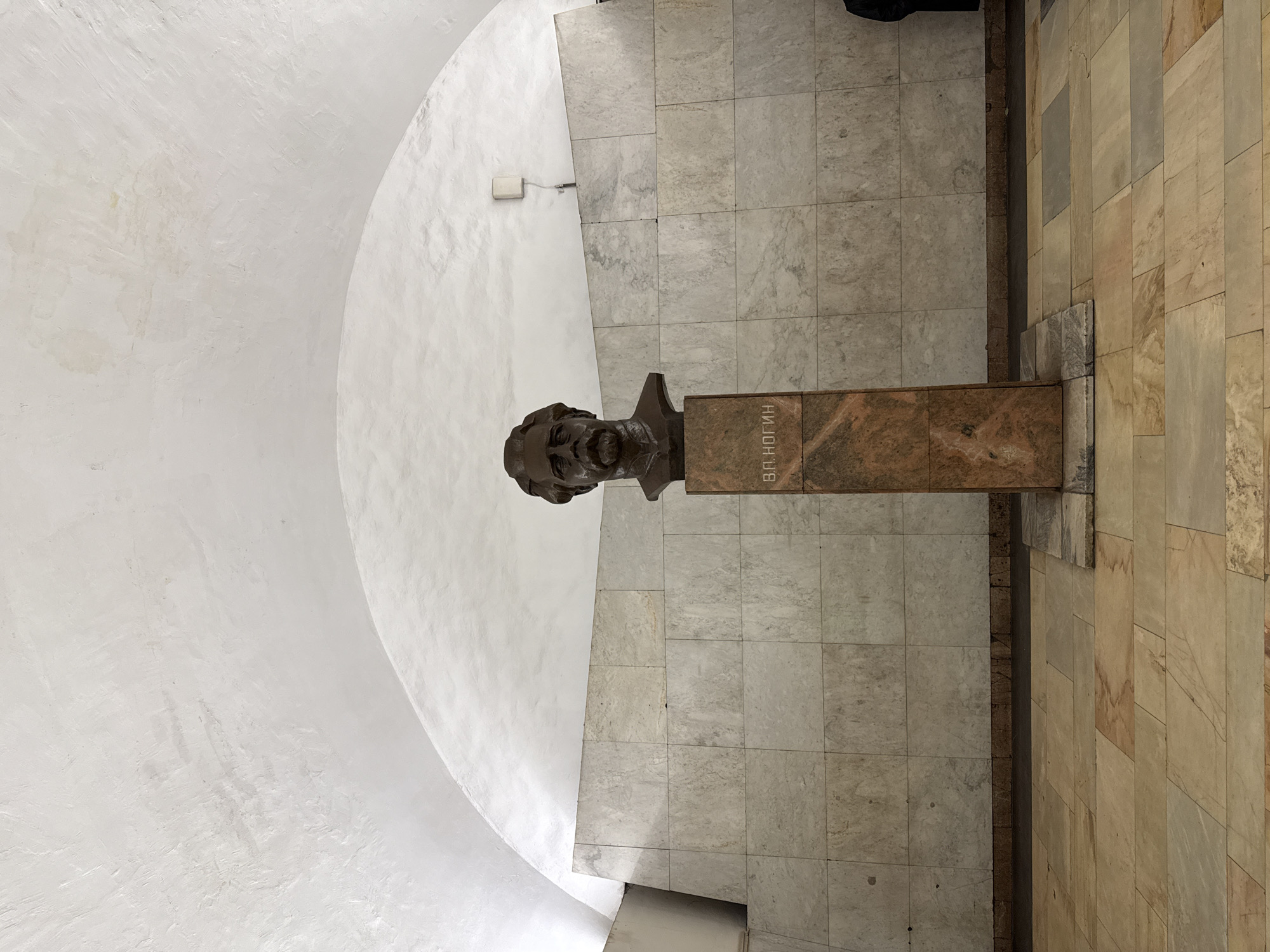 The image shows a bronze bust of Vladimir Ilyich Lenin, in a metro station of Unknown, Unknown. The bust is placed on a brown marble plinth. The station walls are tiled. The ceiling has a curved section.