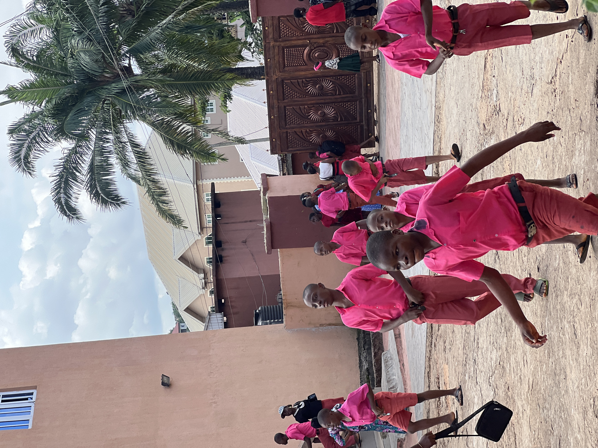 The image depicts a group of young people, predominantly boys wearing matching pink shirts and red shorts, in an outdoor setting. The architecture suggests an urban or semi-urban environment, possibly a school or a community center, with a prominent building featuring a light-colored stucco facade and a blue-paned window on the left. In the background, other buildings with beige roofs and some palm trees are visible. A large, ornate wooden gate stands on the right side of the image. Some individuals are in motion, walking or looking around, indicating a gathering or a school activity. The ground appears to be paved or a dirt-like surface. The overall scene is brightly lit by daylight under a cloudy sky. It is highly probable that this image was taken in the city of Nnewi, Nigeria, given the context of typical school attire and architectural styles found in the region.