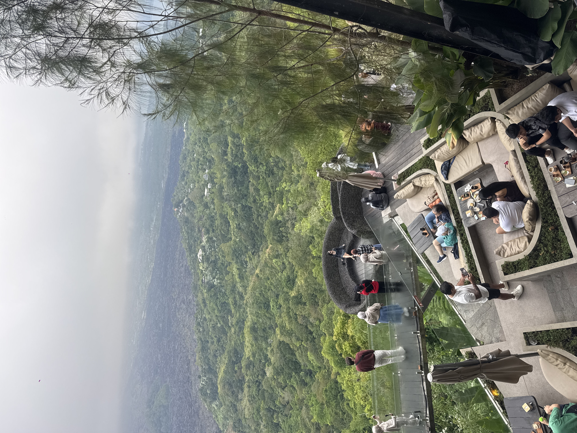 The image depicts an outdoor seating area of a restaurant or cafe perched on a cliffside, offering a panoramic view of a lush, green valley and a misty, volcanic landscape in the distance. In the foreground, several groups of people are visible. On a raised wooden deck, a group of individuals are seated on large, curved beige sofas, some with drinks on low tables. A man in a white t-shirt and black shorts stands in the center of this deck, looking at his phone. Further back, on a transparent walkway extending over the cliff edge, a few people are standing, observing the view. One woman in a black dress is seated on a large, woven circular sofa, also overlooking the valley. The overall environment suggests a scenic tourist spot in Kintamani, Indonesia, known for its volcanic caldera views. The weather appears to be overcast, contributing to the misty atmosphere.
