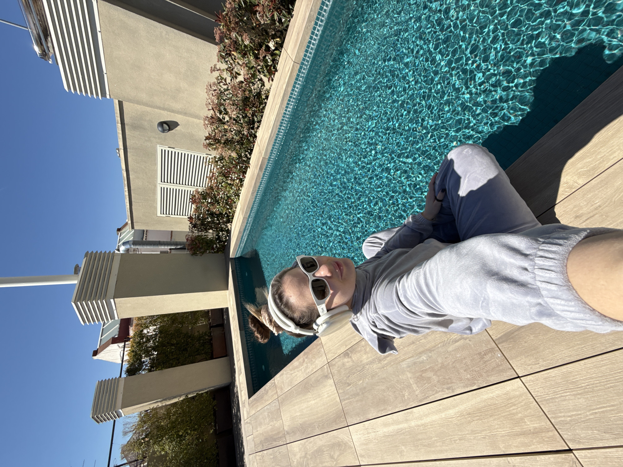 A person is lounging on a tiled deck next to a pool. The individual is wearing sunglasses, headphones, and a gray velour tracksuit. The pool has turquoise water. Behind the pool are trees and a building with architectural details. The sky is clear and blue. The location is the city of Barcelona, Spain.