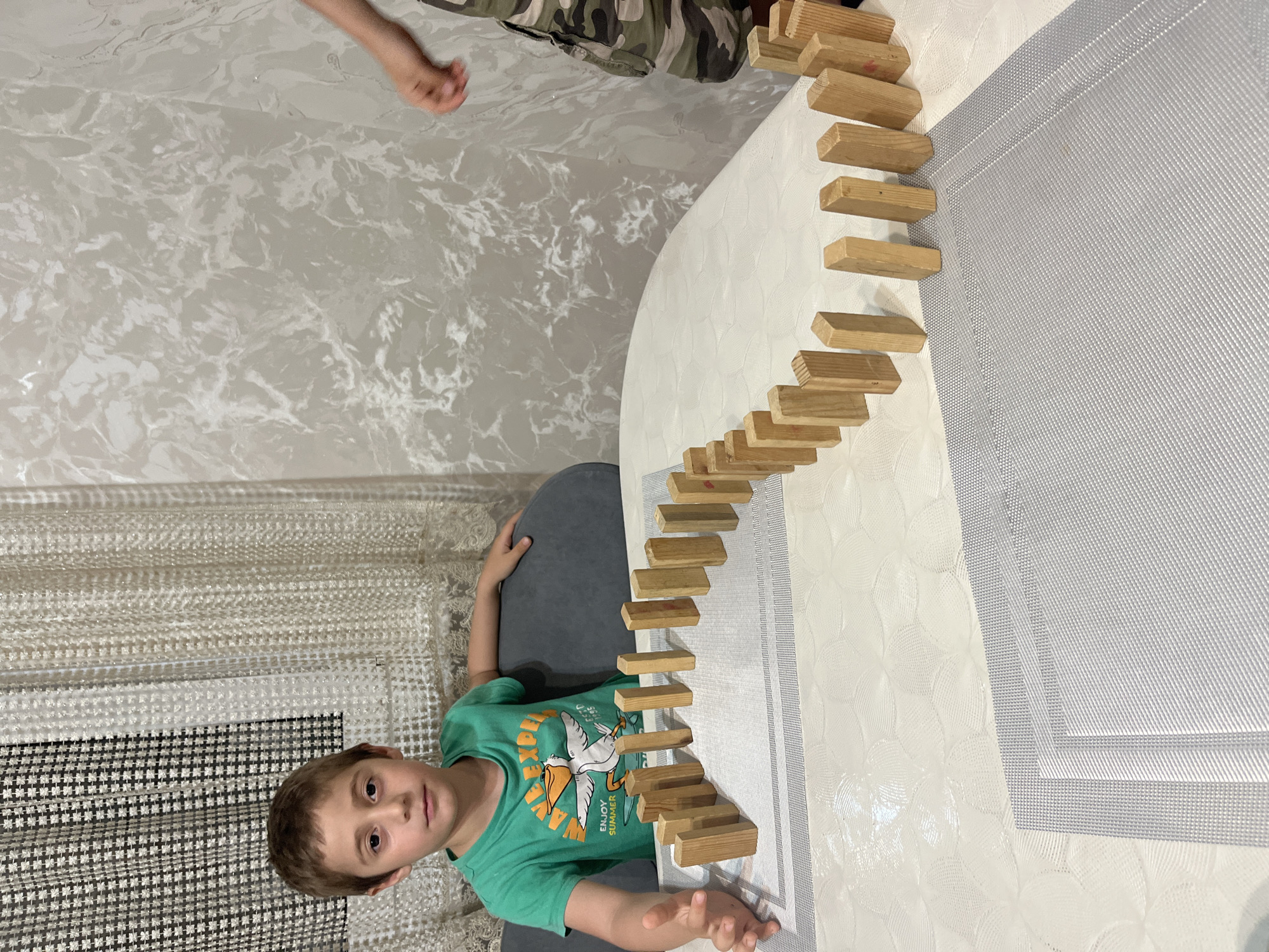 The image depicts a young boy, likely between 5 and 7 years old, sitting at a table. He is wearing a green t-shirt with a graphic print of a pelican and the words "ENJOY SUMMER." His left arm is extended and slightly blurred, suggesting he might be in motion or about to interact with the dominoes. The table is covered with a white tablecloth and placemats. A line of wooden dominoes is set up on the table, forming a curving path. In the background, a second person, partially visible and wearing camouflage shorts, has their right arm extended towards the dominoes, implying they may also be involved in the activity. The room is furnished with curtains and patterned wallpaper. The context of Kaspiysk, Russia, is not ascertainable from the visual information provided in the image.
