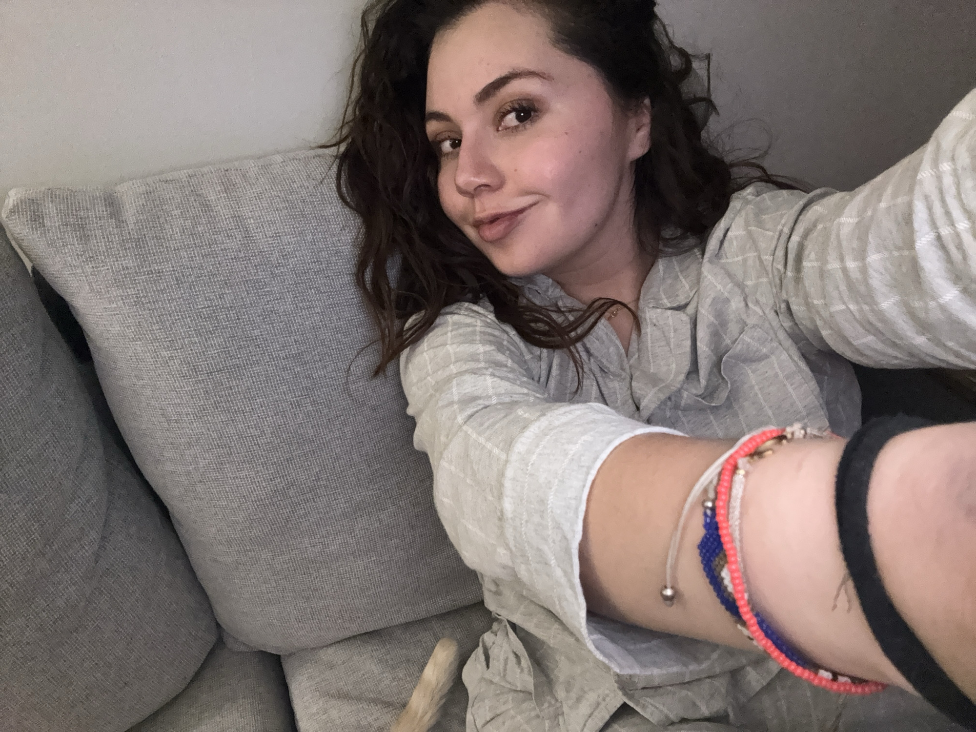 A selfie depicts a woman with curly brown hair, wearing a light gray and white plaid button-down shirt, lying on a gray couch with a gray pillow behind her. She is smiling slightly at the camera. Her arm extends toward the camera, revealing a series of bracelets on her wrist: a white cord bracelet, a blue and white beaded bracelet, a red and white beaded bracelet, and a black band. Part of what appears to be a cat's tail is visible near her waist, suggesting a pet is nearby. The environment is indistinct, with a simple, light-colored wall in the background. There is no information to support the location as Mexico City, Mexico.