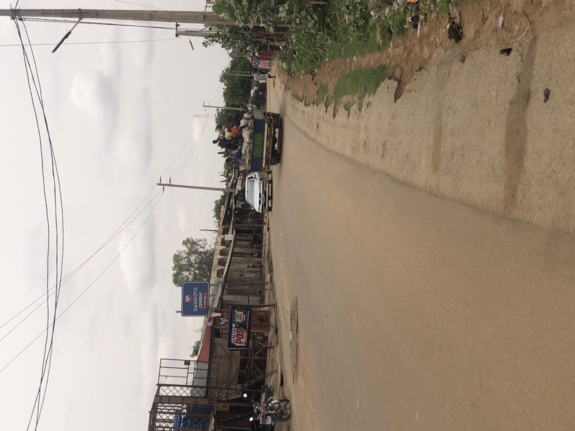 The image shows a street scene in Hanwa, Nigeria. A truck carrying people and goods is traveling on the road. A white car follows behind. The road is paved. On the left side of the road are buildings, including shops, and signs for "Dangote Cement" and "Abtech POS". A motorcycle is parked near one of the buildings. Overhead power lines run above the street. The sky is overcast. Vegetation grows along the roadside on the right. The scene suggests a bustling commercial area.