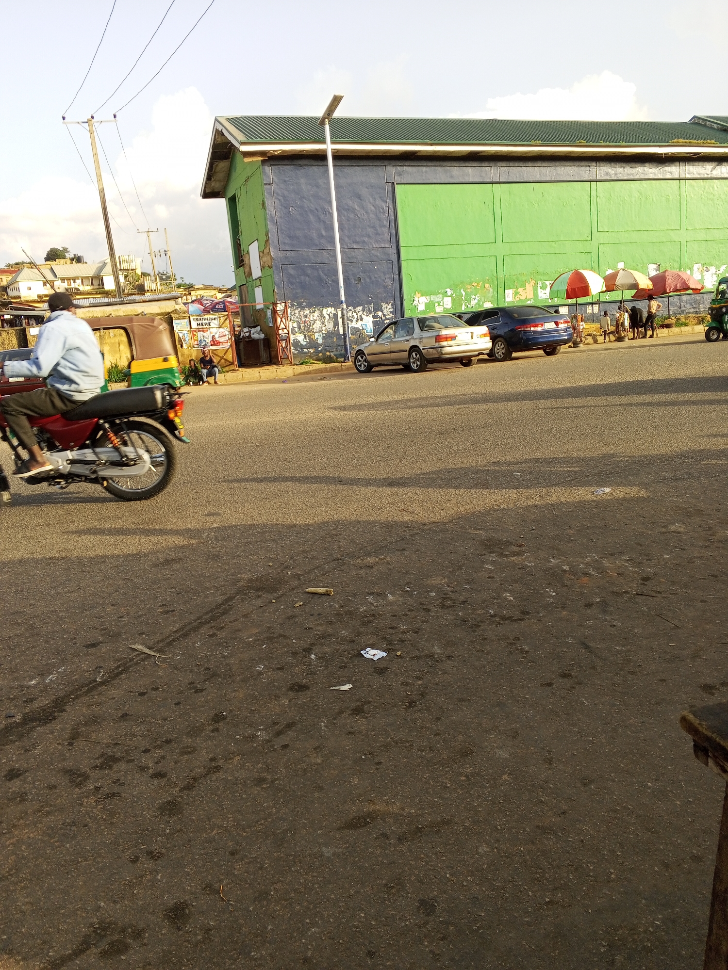 The image captures a street scene in Kabong, Nigeria, under a bright sky, suggesting it is daytime. In the foreground, a person rides a motorcycle, their light blue shirt and dark pants clearly visible. The motorcycle itself is a prominent object, a red and silver model with a black seat. To the left of the motorcycle, a portion of a green and yellow auto-rickshaw is visible, suggesting modes of transportation common in the area.
In the background, a large building with a green roof and walls divided into blue and green sections dominates the scene. This building appears to be a commercial establishment, possibly a market or a shop, with advertisements and posters affixed to its walls. Several cars are parked along the street in front of the building, including a silver sedan and a dark blue sedan.
A group of people are gathered near the building, some standing and others sitting. A few colorful umbrellas are set up, providing shade. The presence of the umbrellas and the general atmosphere suggests a bustling marketplace or a gathering point. The road surface is asphalt, with some debris and oil stains visible, indicating normal street wear and tear. The overall impression is one of a typical day in a busy urban environment in Nigeria. There are no discernible strong emotions evident, but the scene is lively and active. Visible text includes posters on the building, which appear to be advertisements, but the specific content is not legible.