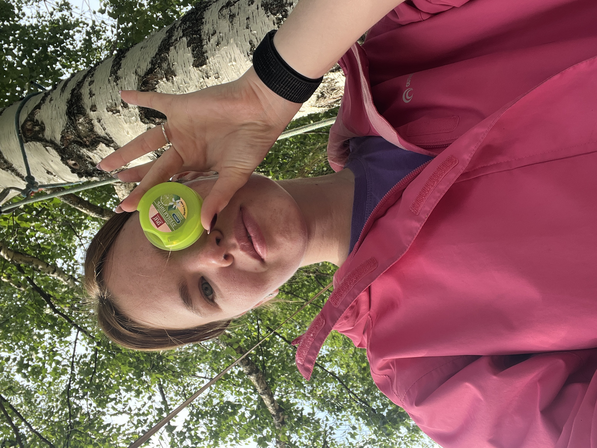 A person, possibly a woman, is shown from a low-angle perspective, looking up towards the camera. They are wearing a bright pink hooded jacket over a purple shirt. A black wristband is visible on their left wrist. In their right hand, they hold a small, round, green container with a label. The person's face is partially obscured by the container.
The background consists of a large birch tree with white bark and dark markings. Lush green leaves and branches from the tree, as well as other trees, fill the upper portion of the frame, suggesting an outdoor setting in a wooded area. The lighting appears to be natural daylight, likely daytime, with dappled light filtering through the leaves. The overall mood seems relaxed and casual.
The label on the green container is partially visible and appears to be in Cyrillic script. Text that can be identified includes "СПЛАТ" (SPLAT) and "ЗУБНАЯ НИТЬ" (dental floss). Other text is too small to be legible.
The location context provided is Sergino, Russia. This suggests the image was taken in a natural environment within that region. The image appears to be a selfie or a candid shot taken outdoors.