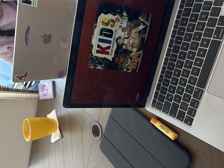 Two open Apple laptops sit on a table. The first laptop is silver and covered in stickers, featuring an Apple logo. The second laptop shows the album "K.I.D.S." by Mac Miller on the screen, with the keyboard visible. A yellow cup rests on a napkin to the side. A purple object and a dark gray tablet case are also present. A tube of lip balm sits near the keyboard.