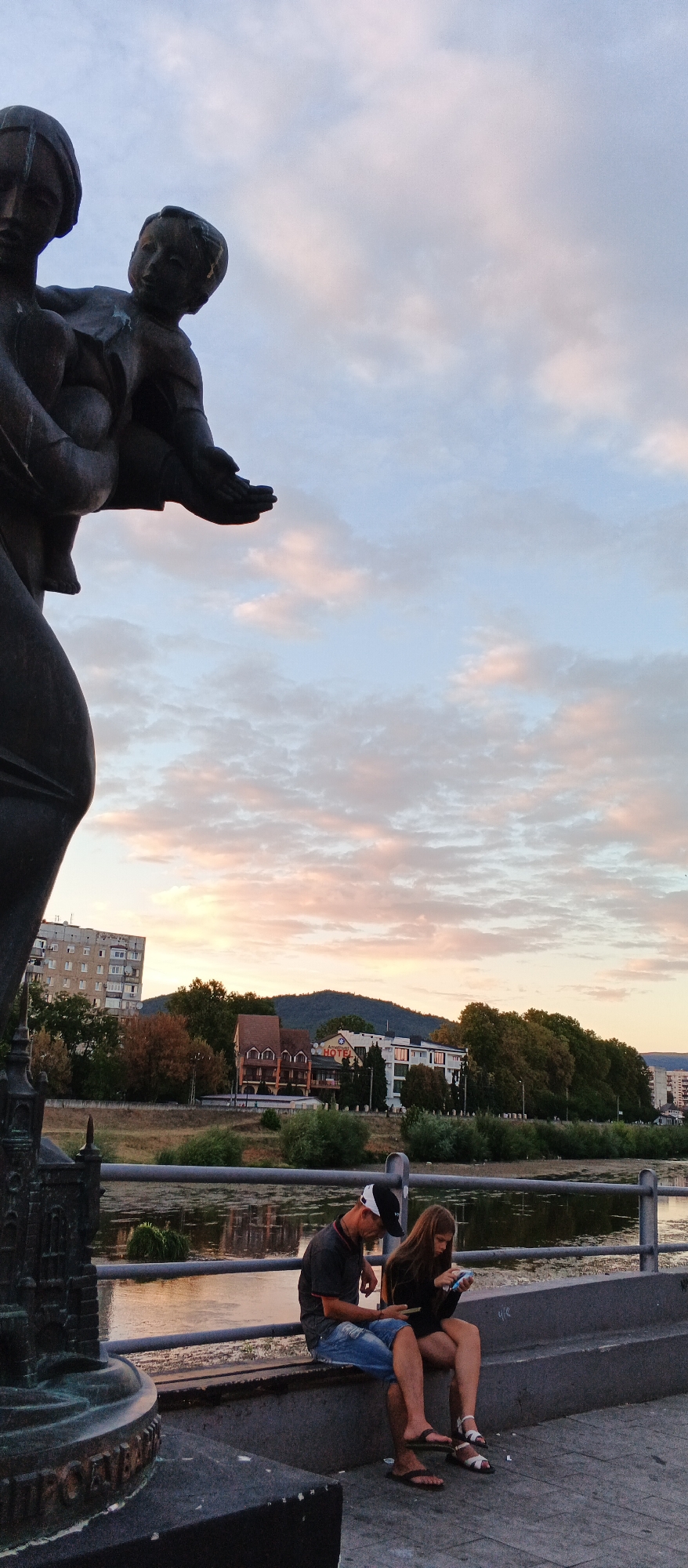 The image depicts a section of the embankment in Mukachevo, Ukraine, during a soft twilight. In the foreground, a dark bronze statue, likely depicting a mother and child, is partially visible on the left. The statue is a monument with intricate details, including architectural elements at its base. A man and a woman are seated on a concrete bench in the mid-ground, facing away from the camera and engrossed in their mobile phones. The man wears a dark t-shirt, denim shorts, and a baseball cap. The woman has long, brown hair and wears a dark top and shorts. They are seated close together, but their interaction is solely with their devices. Behind them, a calm river reflects the muted colors of the sky. The embankment is lined with a metal railing. The background features a treeline, a few buildings including a hotel with signage, and a gentle hill or mountain range. The sky is filled with wispy clouds tinged with pastel hues of pink and orange from the setting sun. The overall atmosphere is tranquil and reflective.