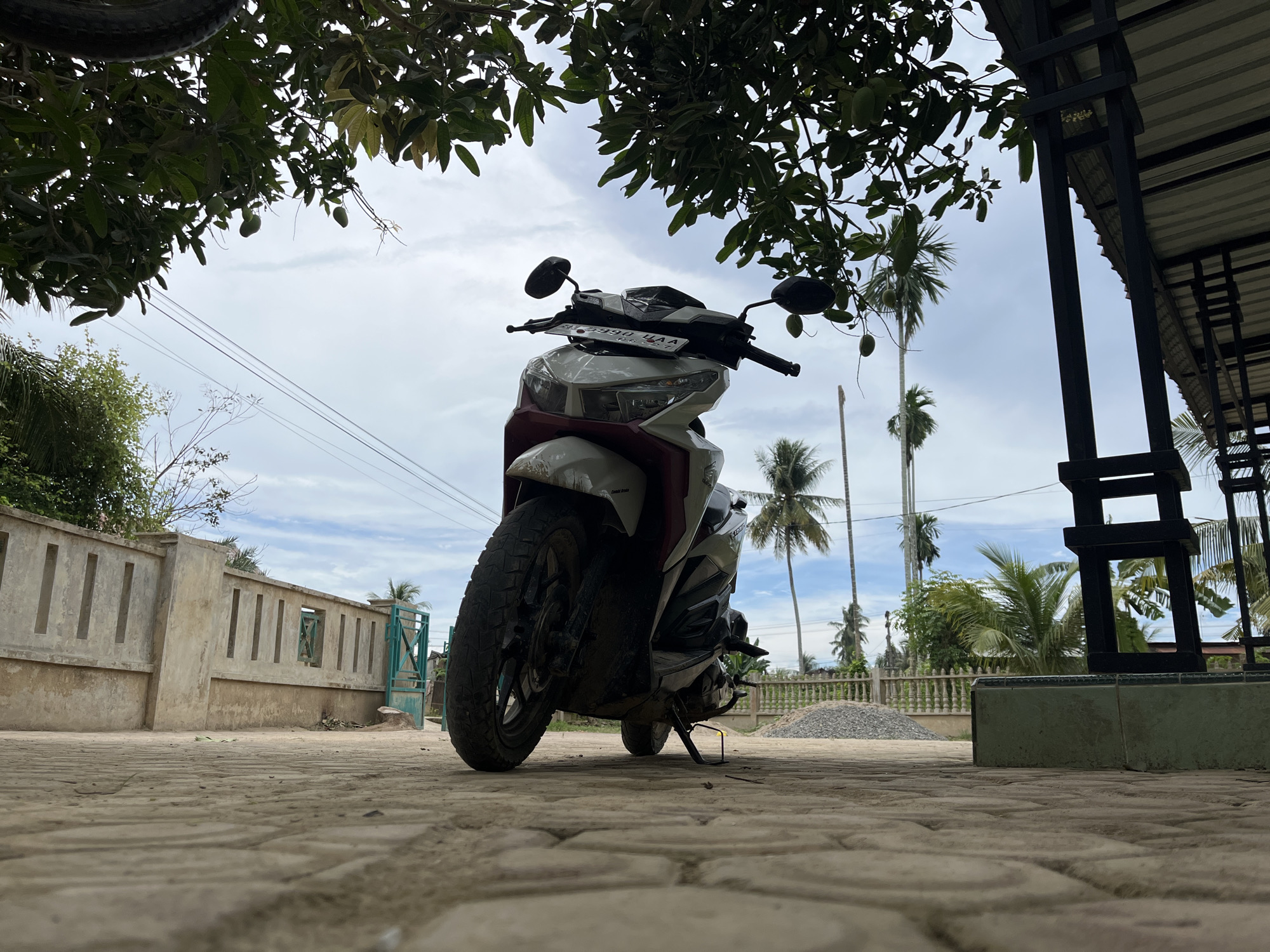 An outdoor scene shows a white and maroon scooter parked on a cobblestone patio. The scooter is viewed from a low angle, with its front wheel prominently featured. Behind the scooter, a concrete wall with rectangular openings runs across the frame, and a teal metal gate is visible. Palm trees and other foliage are scattered throughout the background, with a cloudy sky overhead. A portion of a roof structure with black supports is visible on the right side of the image. The setting appears to be a residential or public outdoor space in Benuaraja, Indonesia.