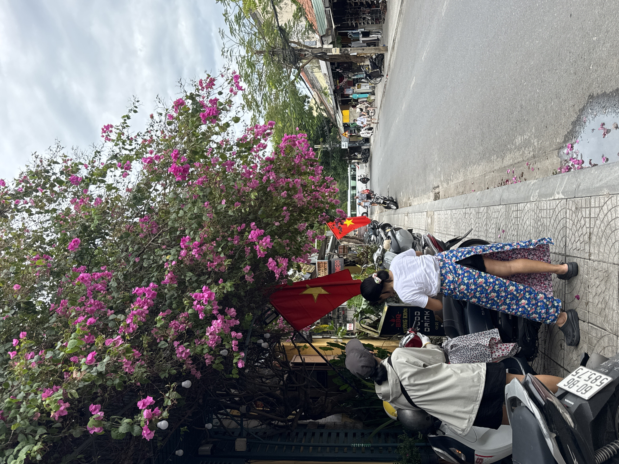 The photograph shows a street scene in Hội An Ward, Vietnam. Two individuals are positioned near parked motorbikes on the sidewalk. One person is wearing a white shirt and a floral-patterned skirt; the other is wearing a gray jacket. There are Vietnamese flags displayed nearby. A large bougainvillea bush with vibrant pink flowers is also prominent. The street leads to shops in the distance.