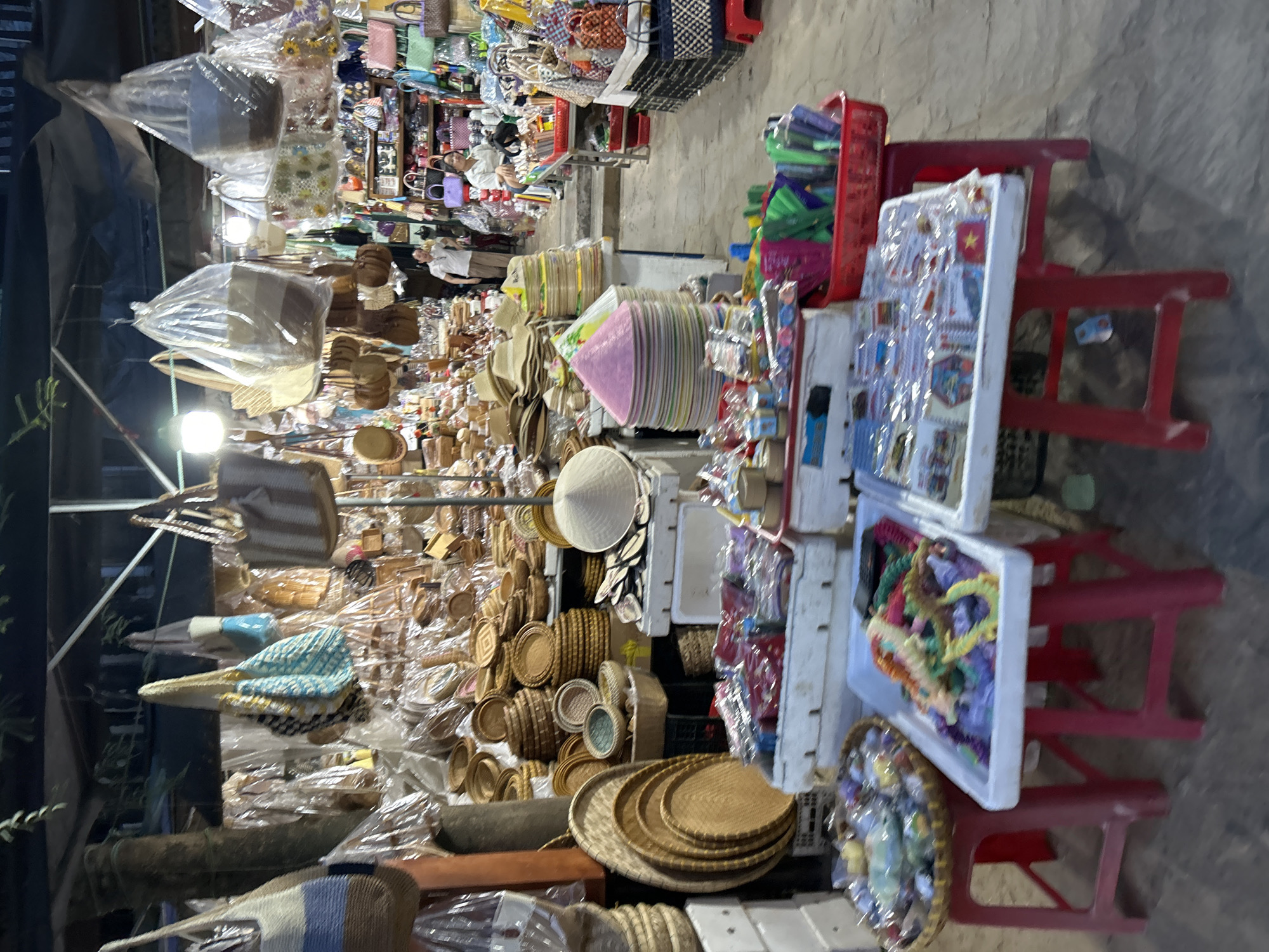 The image depicts a busy marketplace stall, likely in Hội An Ward, Vietnam, characterized by an abundance of handcrafted goods. In the foreground, a red plastic stool supports a woven tray filled with various colorful beads and necklaces. Adjacent to it, another red plastic stool holds a white rectangular tray overflowing with similar colorful beaded jewelry. Further to the right, a third red stool displays a white rectangular tray covered in plastic, revealing a collection of small, packaged items, possibly souvenirs.
Behind these foreground displays, more stalls are visible, laden with an assortment of items. Stacks of woven hats and baskets are prominent, as are numerous bags of various designs and materials, some hanging from the ceiling and others displayed on shelves. The overall impression is of a vibrant and crowded market, with goods neatly arranged for sale. In the mid-ground, two individuals are partially visible, one appearing to be a vendor in a white shirt and the other a woman in a white shirt. The lighting suggests it might be evening, with artificial lights illuminating the scene. The ground appears to be concrete.
