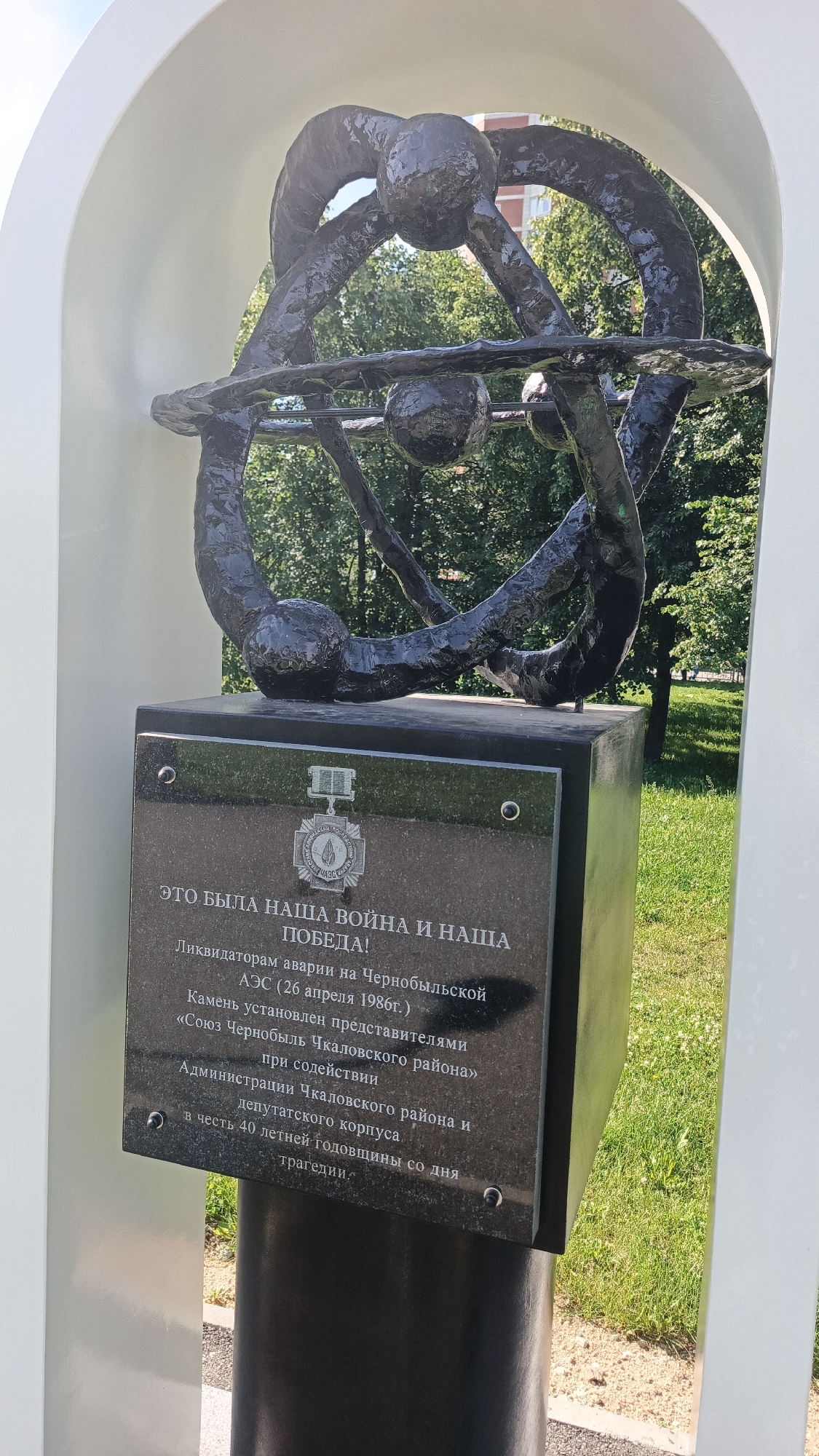 The image shows a monument located in Yekaterinburg, Russia, dedicated to the liquidators of the Chernobyl disaster. The monument features a black granite plaque with Cyrillic text and an emblem. Above the plaque, a dark, abstract sculpture resembling an orrery or atomic model is visible, set within a white arch. The background consists of green grass and trees, suggesting an outdoor setting on a sunny day. The plaque's inscription commemorates the Chernobyl disaster of April 26, 1986, and the 40th anniversary of the tragedy. It also mentions the "Soyuz Chernobyl" (Union of Chernobyl) organization and the Chkalovsky district administration as participants in the monument's establishment.
