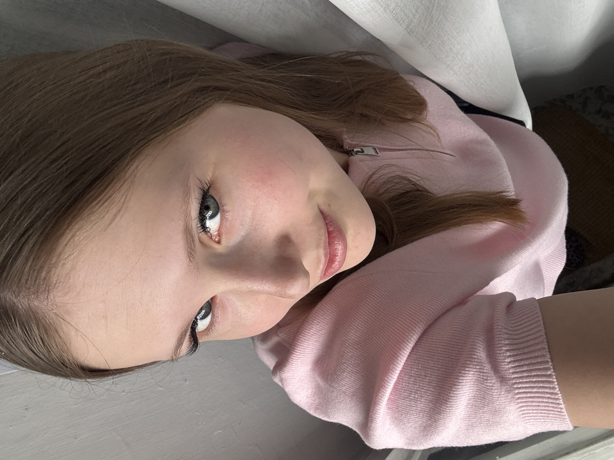 A young woman with light brown hair and blue eyes is facing the camera. She is wearing a light pink sweater. The background includes a gray surface and a white curtain. The location is not identifiable as Chaikovsky, Russia, based on the visual information provided.