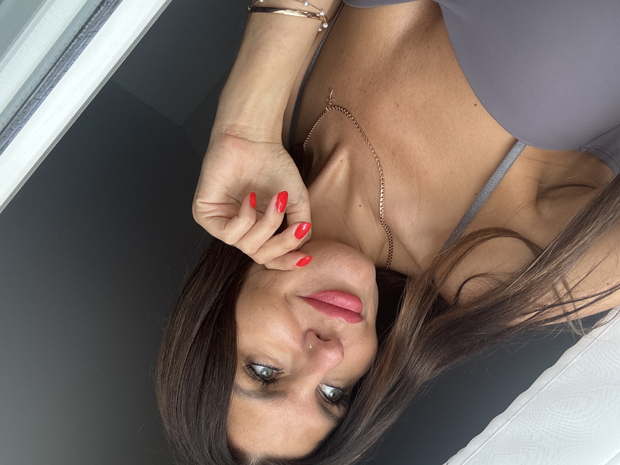 The media file is a close-up, vertically oriented photograph of a woman. She is the sole subject and is positioned centrally in the frame, with her head tilted and her gaze directed slightly off-camera. Her facial features are prominent, including green eyes, full lips with pink lipstick, and dark brown hair. She is wearing a gray top.
Notable details include a delicate gold necklace with a small pendant resting on her collarbone, and a bracelet with pearls on her left wrist. Her fingernails are painted a vibrant red. She has a small nose stud. One of her hands is brought up to her face, with her fingers resting near her mouth and cheek.
The background is divided, with a dark, out-of-focus area on the left and a bright, textured surface on the right, possibly a window blind or curtain. The lighting suggests it might be daytime, as there is bright light coming from the right side of the frame. The overall mood appears relaxed and possibly contemplative. There is no visible text in the image. The location context provided is Chaikovsky, Russia, but there are no direct visual cues within the image to confirm this.
