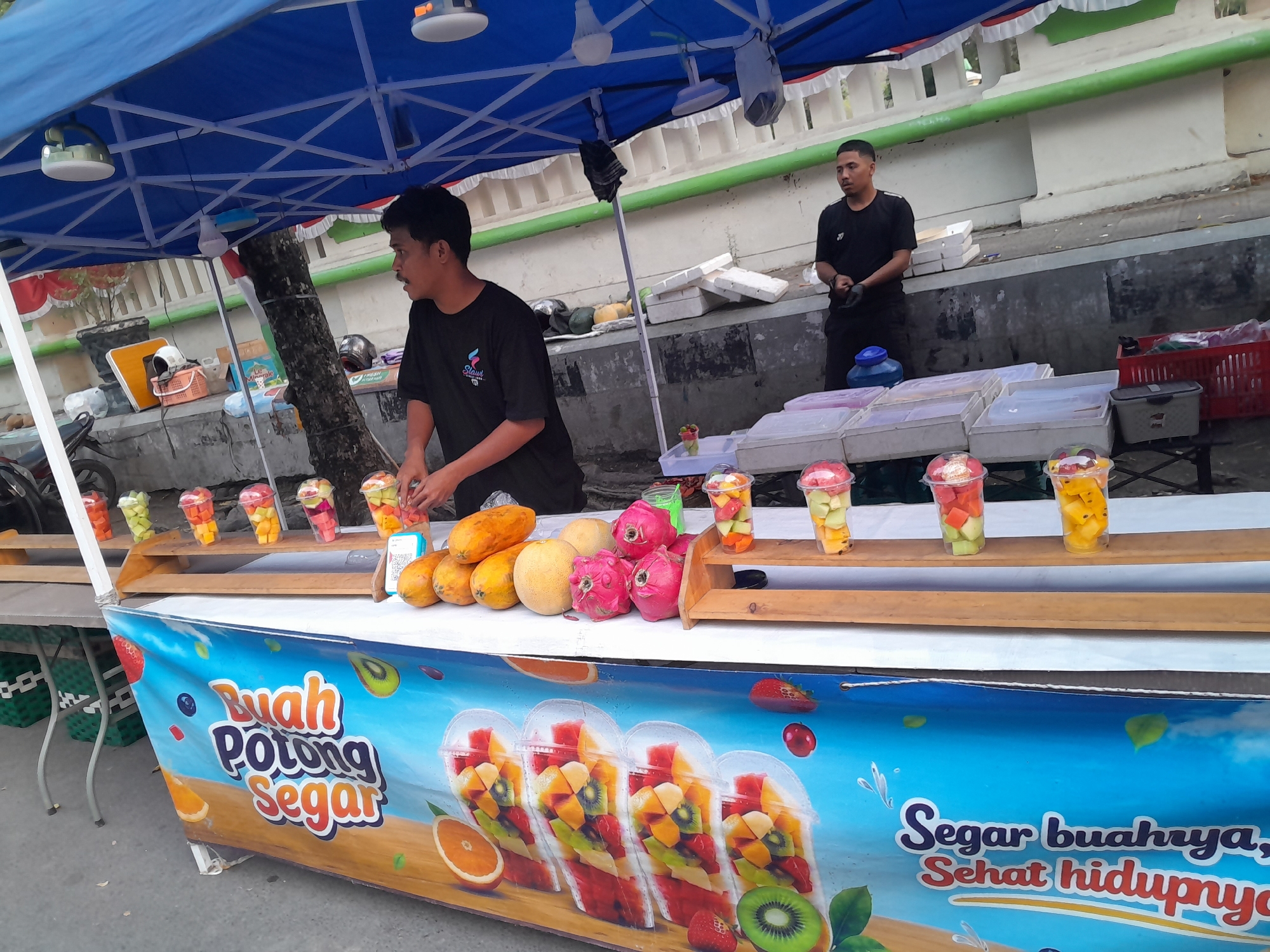 This is a medium shot photograph taken outdoors in Slawi, Indonesia, depicting a fruit stand. Two men are present. The man on the left, wearing a black t-shirt with a colorful logo, is actively preparing fruit. He stands behind a counter laden with various fruits, including papayas, melons, and dragon fruit. Various cups filled with chopped fruit are displayed on a wooden shelf above the counter, with at least six cups visible. The man on the right, also in a black t-shirt, stands behind the fruit stand and appears to be observing. The fruit stand is adorned with a blue banner featuring the text "Buah Potong Segar" (Fresh Cut Fruit) and "Segar buahnya, Sehat hidupnya" (Its fruit is fresh, its life is healthy), along with illustrations of fruits. In the foreground, several clear plastic cups filled with fruit salad are arranged, with at least four fully visible. The background shows a wall and some vegetation. The overall scene suggests a typical street food vendor operating in a public space.