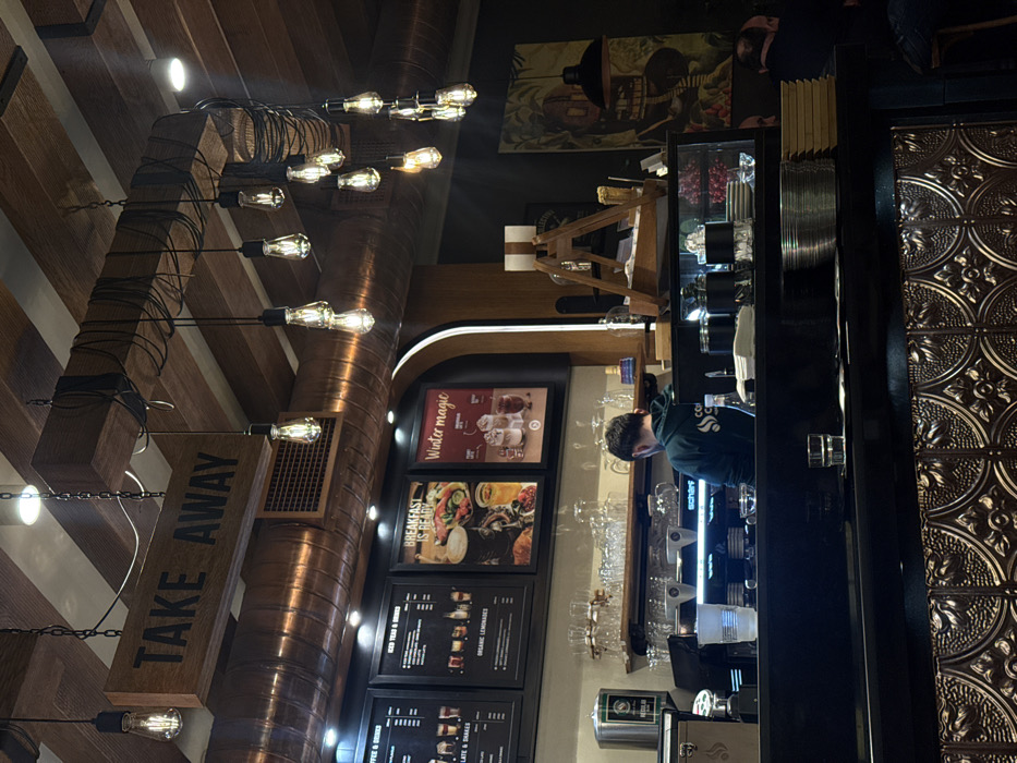 A café interior features a barista behind a counter, surrounded by glassware and coffee equipment. Menu boards display offerings like "Winter Magic" and breakfast options. The ceiling is adorned with exposed wooden beams and vintage-style light bulbs strung along cables. A "TAKE AWAY" sign hangs from one of the beams. A decorative tile pattern covers a section of the wall. A painting is on the upper back wall.