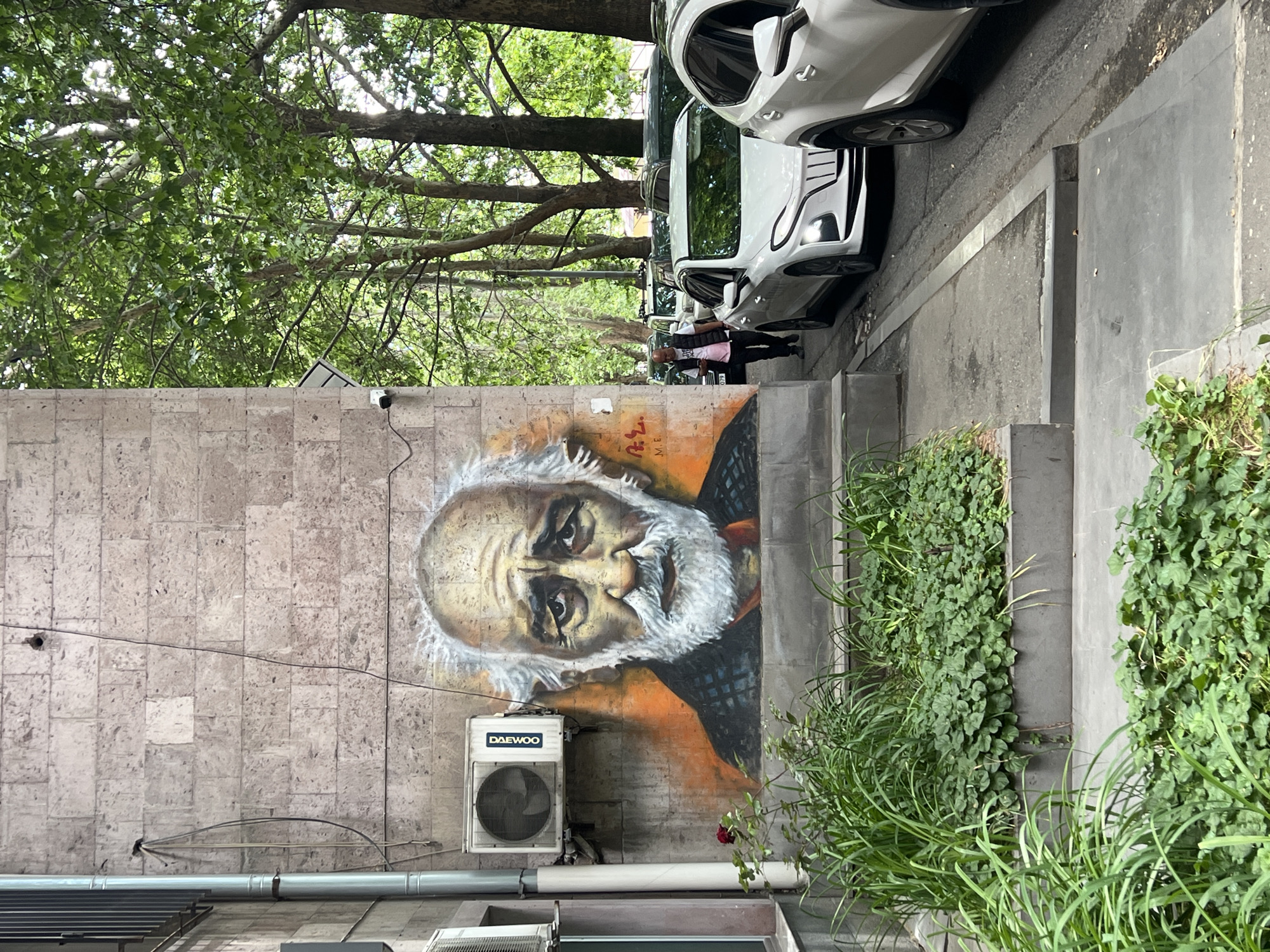 The image shows a street scene in an urban location. A mural of an elderly man with a white beard is painted on a textured stone wall, with a Daewoo air conditioning unit installed below. A metal pipe runs horizontally across the wall beneath the AC unit. The surrounding vegetation includes green bushes and trees, partially obscuring the mural. Several white cars are parked along the street adjacent to the wall. People walk on the sidewalk past the cars. The exact location is unknown.