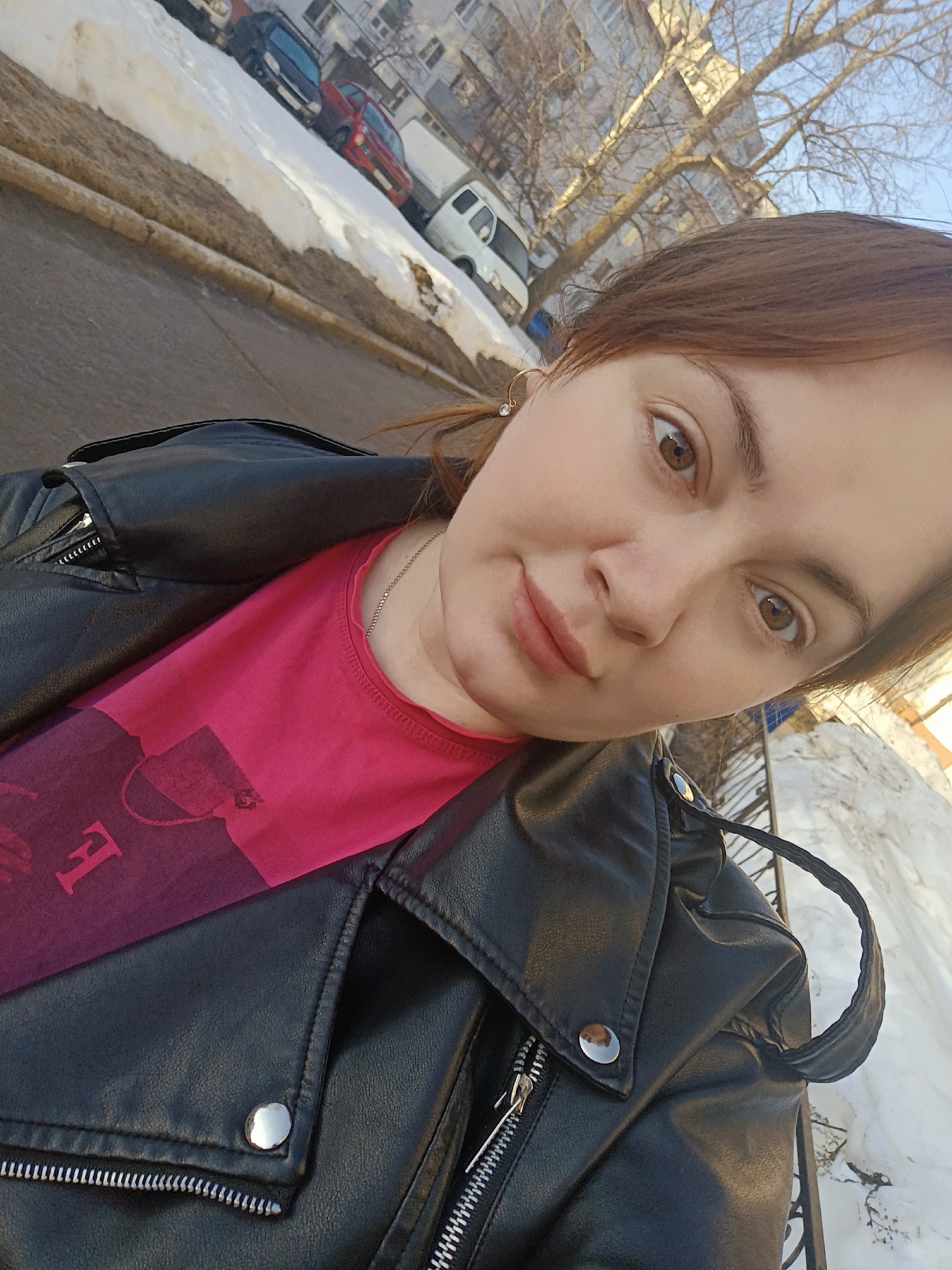 A woman is taking a selfie on a city street in Chaikovsky, Russia. She is wearing a black leather jacket over a pink t-shirt. Patches of melting snow are visible along the side of the road. In the background, there is a street with parked cars and buildings, including a van and a red car. Bare trees are visible in front of the buildings. The lighting suggests it is daytime.