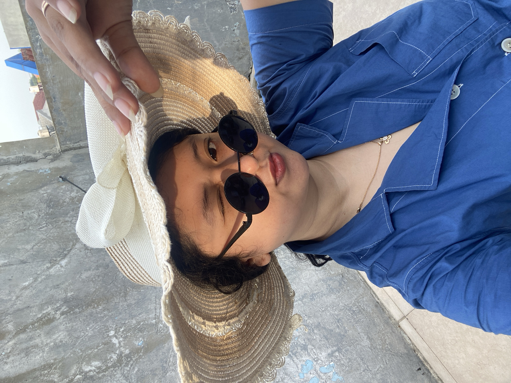 A woman wearing a blue button-down shirt, sunglasses, and a straw hat poses for a selfie outdoors in Gelam, Indonesia. Her hand, with long manicured nails and a ring on her finger, holds the hat. She is lying on a concrete surface that shows signs of wear, possibly a rooftop or patio, with some nearby buildings and structures visible in the background. The lighting suggests it is daytime. She is also wearing a delicate gold chain.