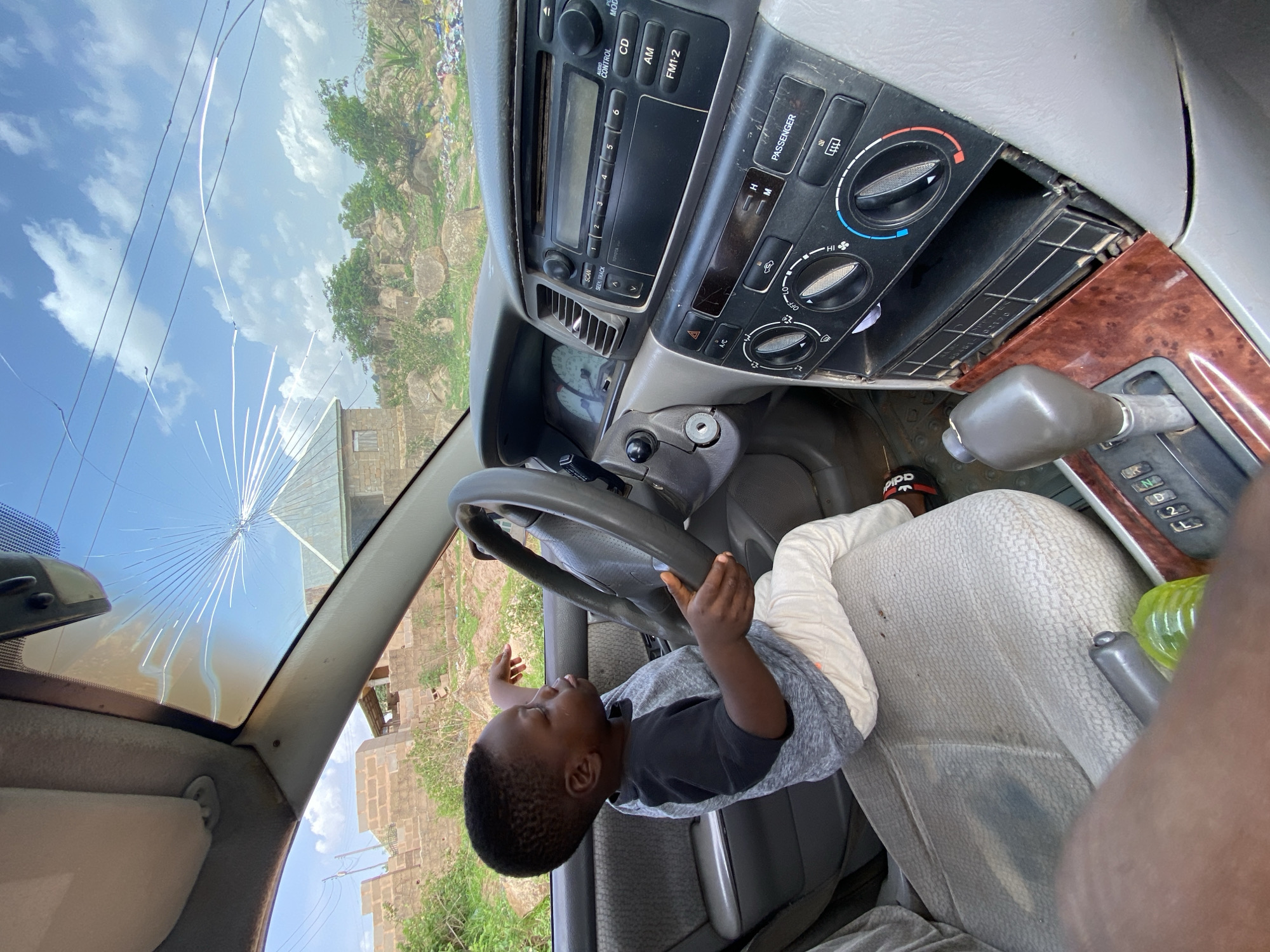 The interior of a car with a cracked windshield. A young child is sitting in the driver's seat, holding the steering wheel. The car's dashboard, radio, and gear shift are visible. Outside the car, vegetation, a blue sky with clouds, and a building are visible. The location is Barakin Akawo, Nigeria.