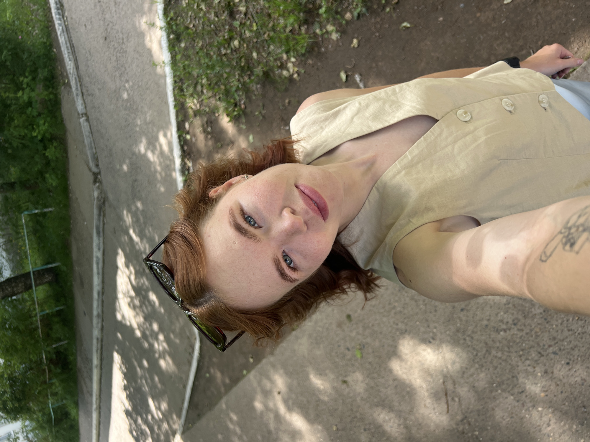 A young Caucasian woman with red hair and light blue eyes is taking a selfie outdoors on a sunny day. She is wearing a light beige sleeveless vest with three buttons and light-colored pants. Sunglasses are perched on top of her head. The background shows a paved area with shadows from trees, a curb, and greenery with fallen leaves. The scene appears to be in a park or residential area, possibly near a sidewalk or road in Chaikovsky, Russia.