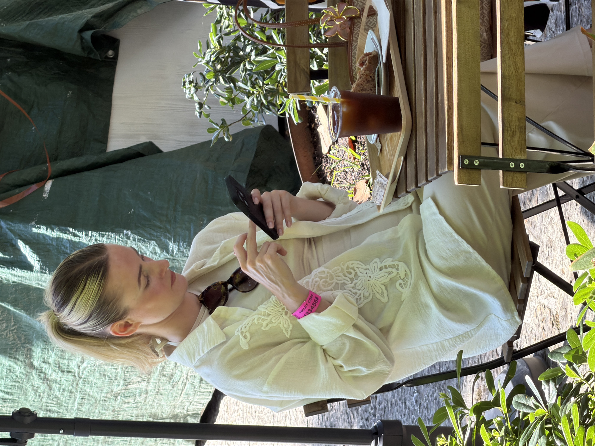 Captured from an overhead perspective, the image portrays a woman seated at a slatted wooden table in what appears to be an outdoor setting, possibly in Bunyola, Spain. The woman is fair-skinned with blonde hair styled in a low ponytail, and she's engaged with her smartphone, held in her left hand. She is dressed in light-colored clothing, including a white shirt with lace detailing and a matching dress. A pink wristband adorns her left wrist, with the words "TOILET CODE" and a number "123456#" printed on it. A pair of sunglasses rests on her shirt, close to her collar.
The table in front of her holds a tray with a beverage in a clear plastic cup, complete with a straw, and what seems to be a pastry. Greenery is also part of the table setting, and foliage is visible on the periphery of the image. A green tarp is seen in the backdrop, providing a shaded area. The seating consists of a metal-framed chair that appears to be part of an outdoor dining set. The ground beneath the chair seems to be paved or made of stone.