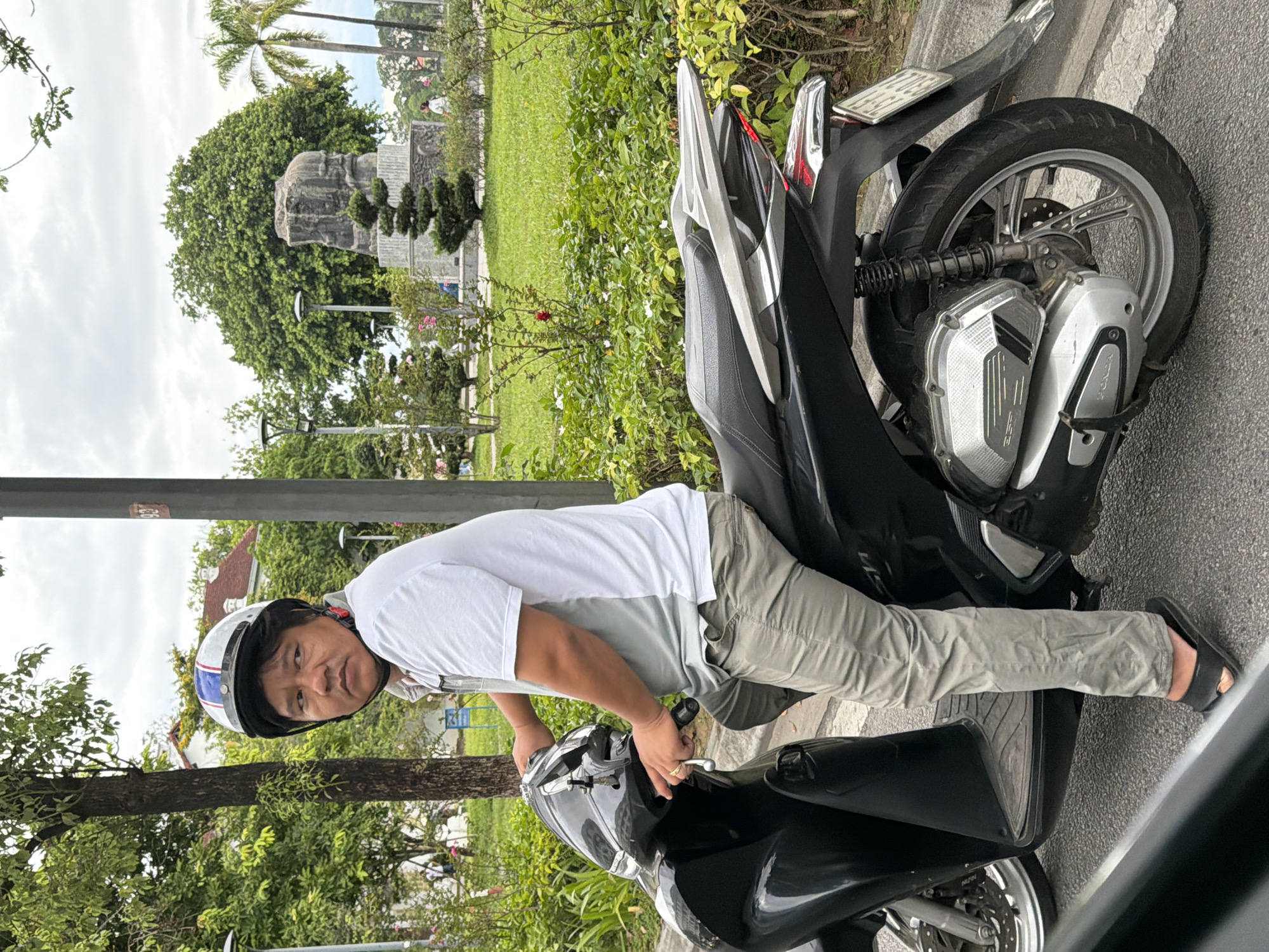 A man wearing a white t-shirt, grey pants, and a helmet is sitting on a black scooter on a paved road. He is looking to his left. The scooter is parked next to a curb and a patch of green grass and bushes. In the background, there are trees, a statue, and buildings. The sky is overcast. The man appears to be in a park or public area in Thuận Hóa Ward, Vietnam.