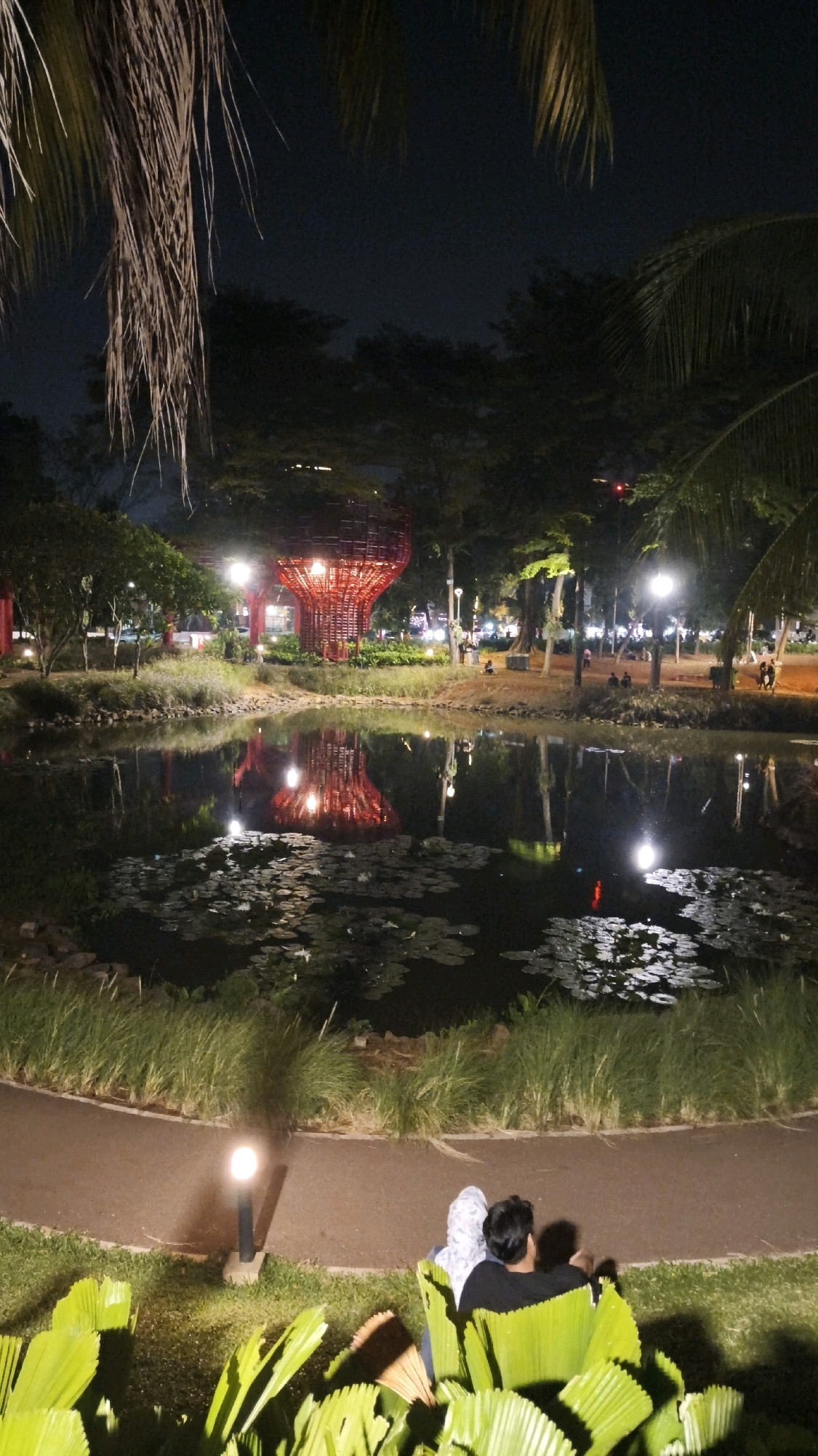 This is a nighttime photograph of the Tebet Eco Park in South Jakarta, Indonesia. The park features a central pond with lily pads, reflecting the ambient lights and a prominent red, abstract sculpture. Several trees surround the pond and sculpture, and pathways are visible. In the foreground, two people are seated on the grass, facing away from the camera, looking towards the pond. The overall scene is illuminated by park lighting, creating a serene atmosphere.
