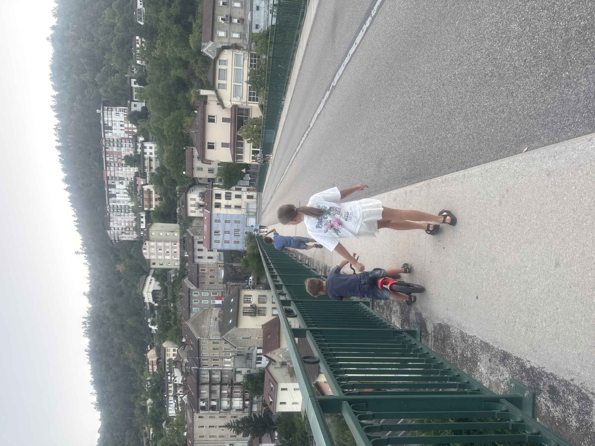 The image shows a girl in a white t-shirt and skirt standing on a bridge, with a boy on a small bike next to her. A third person, possibly a boy, is lying on the bridge in the background. The bridge overlooks a town with many buildings nestled on a hillside covered in trees. The architecture of the buildings suggests an European location, possibly Saint-Claude, France, given the location context. The weather appears overcast, with a hazy sky. The time of day is difficult to determine due to the lack of strong shadows, but it is likely daytime. The overall mood seems relaxed and familial, with children engaged in playful activity. No discernible text is visible on the people or their clothing.