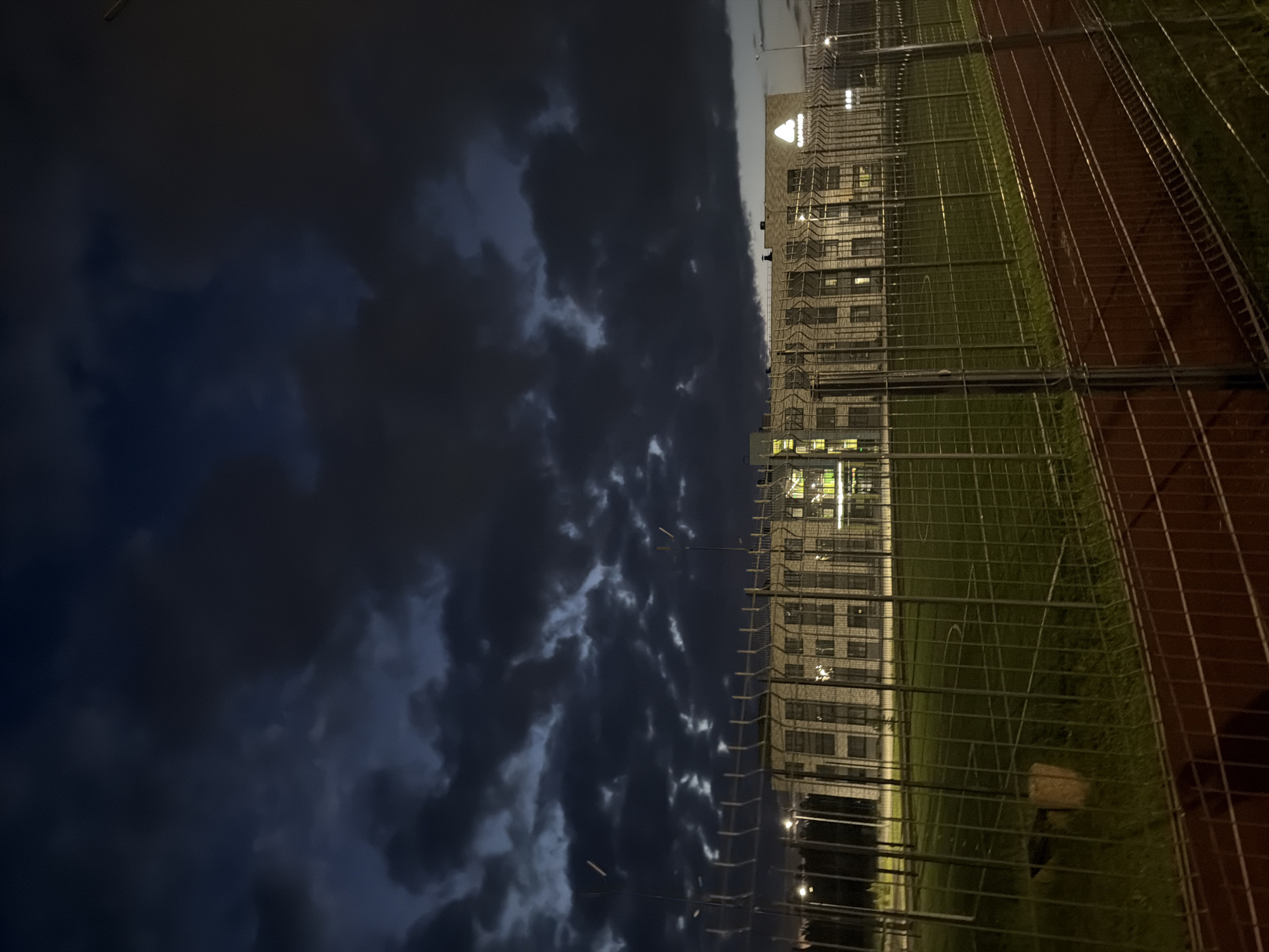 The image captures a wide shot of a building at dusk in Naberezhnye Chelny, Russia, under a cloudy sky. In the foreground, a chain-link fence encloses a grassy area, which appears to be a sports field or park. Beyond the fence, a multi-story building with numerous windows is visible, illuminated from within. The building's facade is constructed of brick-like material. A logo, a white triangle within a circle, is prominently displayed on the building's upper section, accompanied by text that appears to be in Cyrillic. The overall atmosphere is subdued due to the low light conditions.