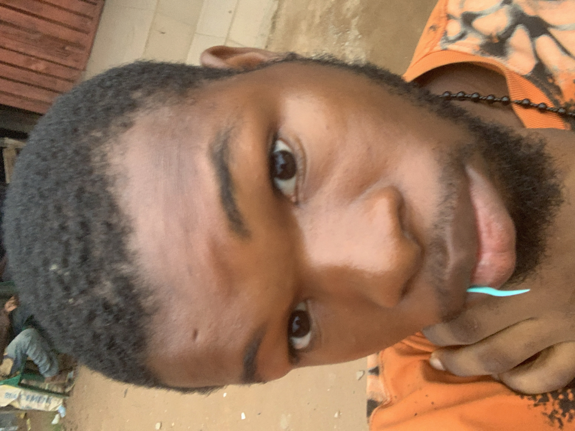 A close-up, vertically oriented photograph captures a young man with dark skin, short curly black hair, and a neatly trimmed beard. He is looking directly into the camera with dark brown eyes and a subtle smile or smirk. A distinctive light blue, thin, curved object, possibly a dental floss pick, is held playfully between his full lips. Around his neck, a black beaded necklace is visible. He is wearing an orange t-shirt with a darker, abstract, possibly tie-dye, pattern on the chest.
The background suggests an outdoor setting in Onitsha, Nigeria, likely during the daytime. To the left, behind his head, is a wooden structure with horizontal planks, resembling a wall or shutter. The ground beneath him appears to be light-colored and uneven, possibly paved or sandy. In the lower left corner, obscured by the subject's shoulder and head, there are faint glimpses of a person in grey trousers and some large bags, possibly cement or grain bags, with some illegible text, indicating a bustling, urban or commercial area. The overall lighting is bright and natural, casting soft shadows.