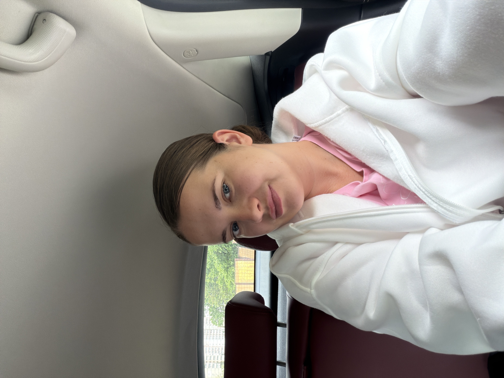 A woman is in a car. She wears a white hooded sweatshirt over a pink top. The car's interior is gray and burgundy. Outside the window, trees and a building are visible. The location is Sochi, Russia, based on the architecture of the building seen outside the window.
