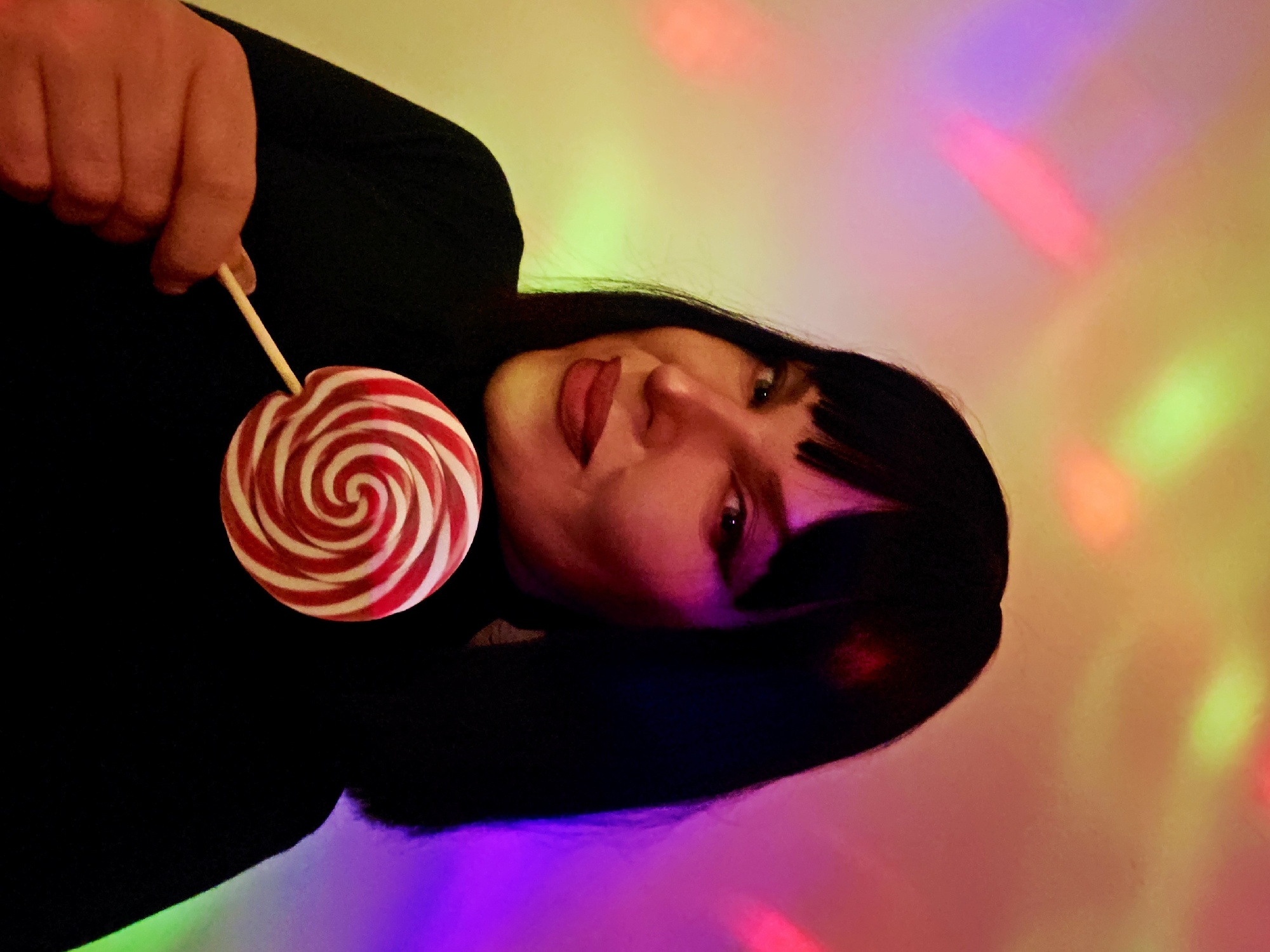 A woman with dark hair is holding a red and white swirl lollipop. The background has colorful lights in various shades of pink, green, and yellow. The scene's location is unknown.