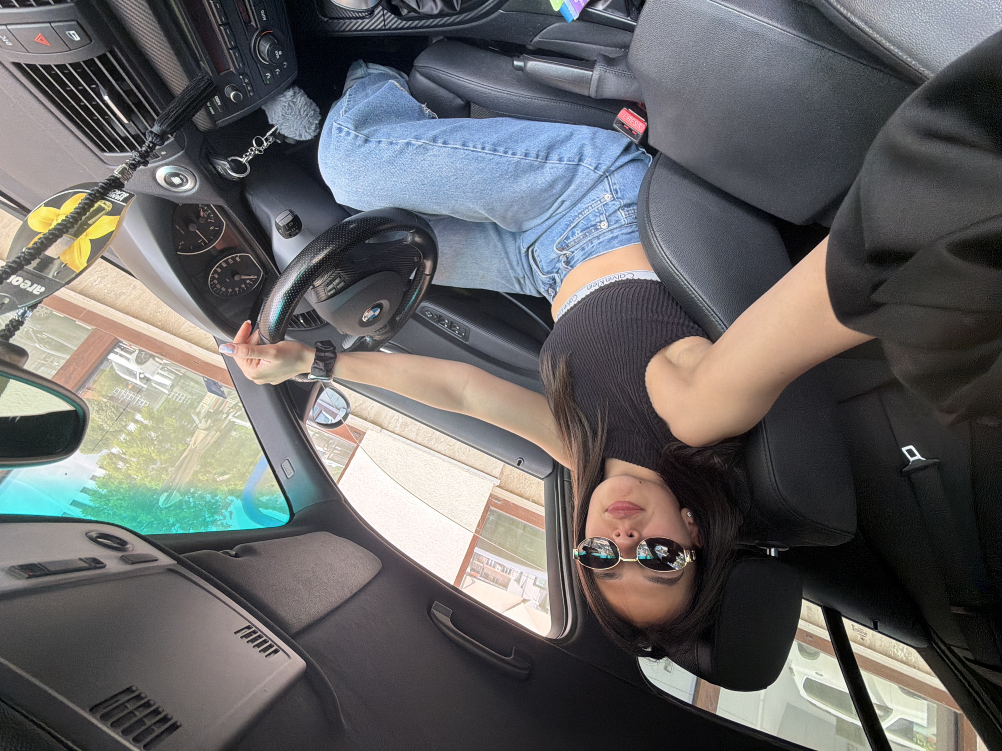 This wide-angle photograph, taken inside a car in Gabrovo, Bulgaria, is rotated 90 degrees clockwise, giving the impression that a young woman is suspended upside down across the front seats. The primary subject is a young woman with long dark hair and reflective sunglasses, looking directly at the camera with a neutral expression. She is wearing a black ribbed crop top, light blue jeans, and a visible "Calvin Klein" waistband. Her left arm extends towards the steering wheel, while her right arm is bent near her head.
The car's interior is detailed, featuring a black carbon fiber-look steering wheel with a visible "BMW" logo. The dashboard includes an instrument cluster with a speedometer and tachometer, as well as air vents. The center console displays an "AUTO" button on the stereo/climate unit and a hazard light button. A key fob with a fluffy grey keychain hangs near the ignition. A black and yellow air freshener with a floral design and partially visible text, including "forea," dangles from the rearview mirror. Black leather seats and a central armrest are also visible. Red "PRESS" and "SOS" buttons are present near the seatbelt buckle area.
Through the car's windows, a bright daytime scene is visible, showing green trees, light-colored buildings, and other vehicles parked in the distance, indicating an urban or suburban setting. The bright light suggests a clear day. The activity is a posed photograph, with the woman casually lying across the front seats for the shot, creating a dynamic and unconventional perspective.