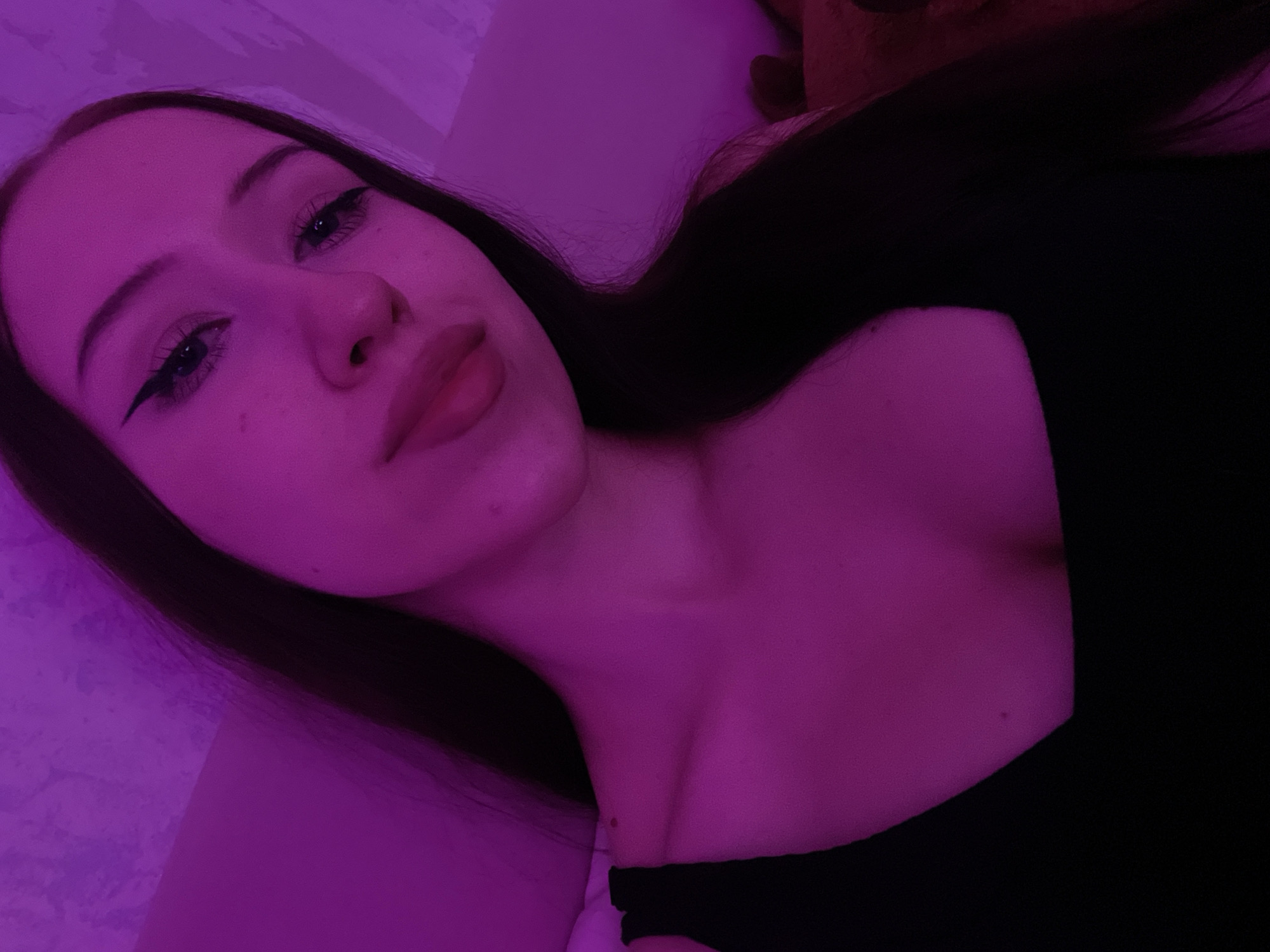 A young woman with long, dark hair is taking a selfie in a dimly lit room with purple ambient lighting. She is wearing a black top with a square neckline. Her makeup includes winged eyeliner and pink lipstick. The background appears to be a wall or soft surface, possibly a bed or couch. There are no discernible landmarks or objects that would indicate a specific location within Votkinsk, Russia.