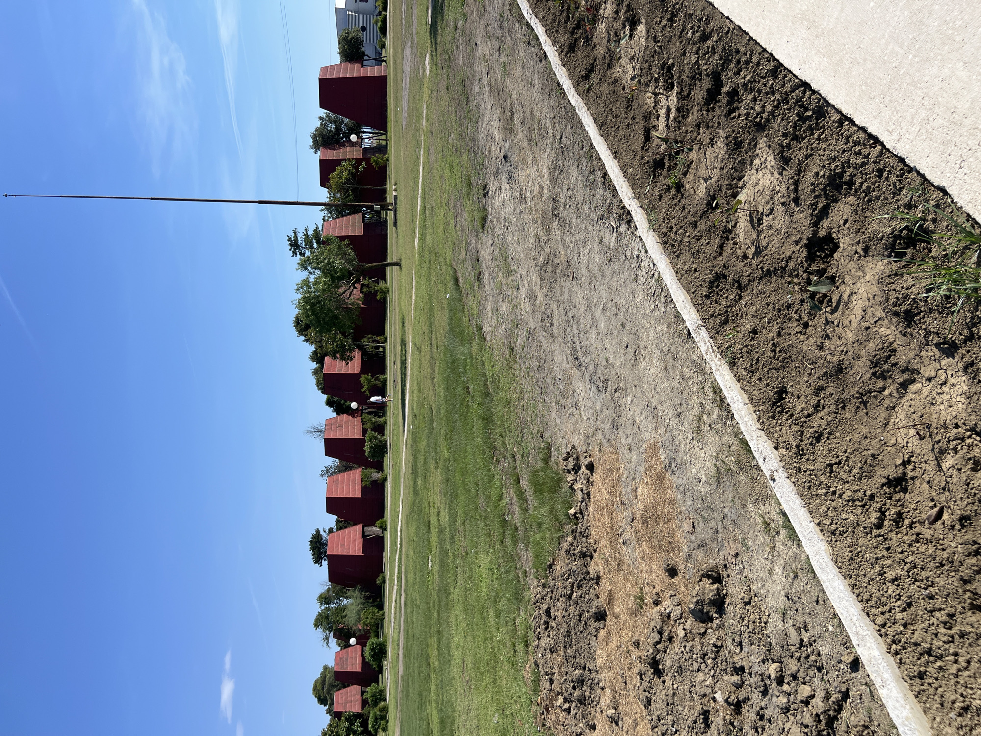 The image depicts a clear blue sky over a landscape in Gornji Štoj - Shtoji i Epërm, Montenegro. In the foreground, a strip of tilled earth is visible, separated by a concrete border from a grassy area. Beyond the grass, a line of red, angular structures, resembling small buildings or pavilions, are arranged in a row. These structures have a distinctive stepped roof design. Trees are interspersed among the red structures, and a tall, thin pole, possibly a flagpole, extends upwards into the sky. The scene appears to be outdoors during the daytime, with bright sunlight casting shadows. The overall impression is one of an open, possibly public or recreational space under construction or preparation for an event, given the freshly tilled soil.