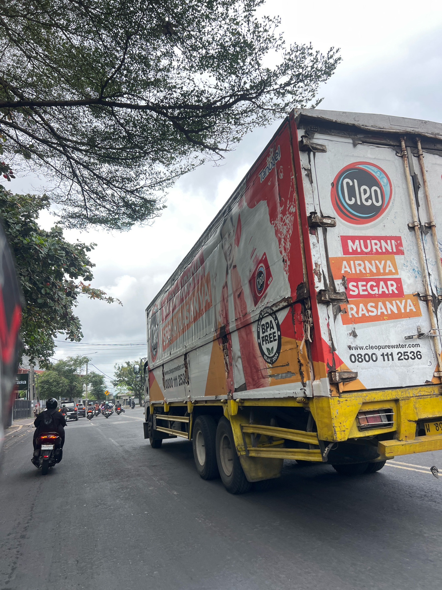 This is a street-level, eye-level photograph taken from a vehicle on a cloudy day in Denpasar, Indonesia, likely on a main thoroughfare. The primary focus is a large, white and red truck carrying a prominent "Cleo" logo, a brand of bottled water. The truck has text on its side which reads "MURNI AIRNYA SEGAR RASANYA," meaning "Pure water, fresh taste." Below this text, a website "www.cleopurewater.com" and a phone number "0800 111 2536" are visible. The truck is yellow on its lower chassis.
In the foreground, the left side of a motorcycle and its rider, wearing a black jacket and helmet, are captured. The motorcycle is moving forward on the asphalt road, which has visible lane markings. Several other motorcycles are also seen on the road ahead, suggesting moderate traffic. A few cars are also present in the distance.
The environment includes trees with dense foliage on the left side of the frame, casting shadows. Buildings, possibly shops or businesses, are visible along the side of the road, with signage like "asa" on one of them. Overhead, telephone or power lines crisscross the sky. The overall impression is of a typical urban scene in Indonesia during overcast weather.