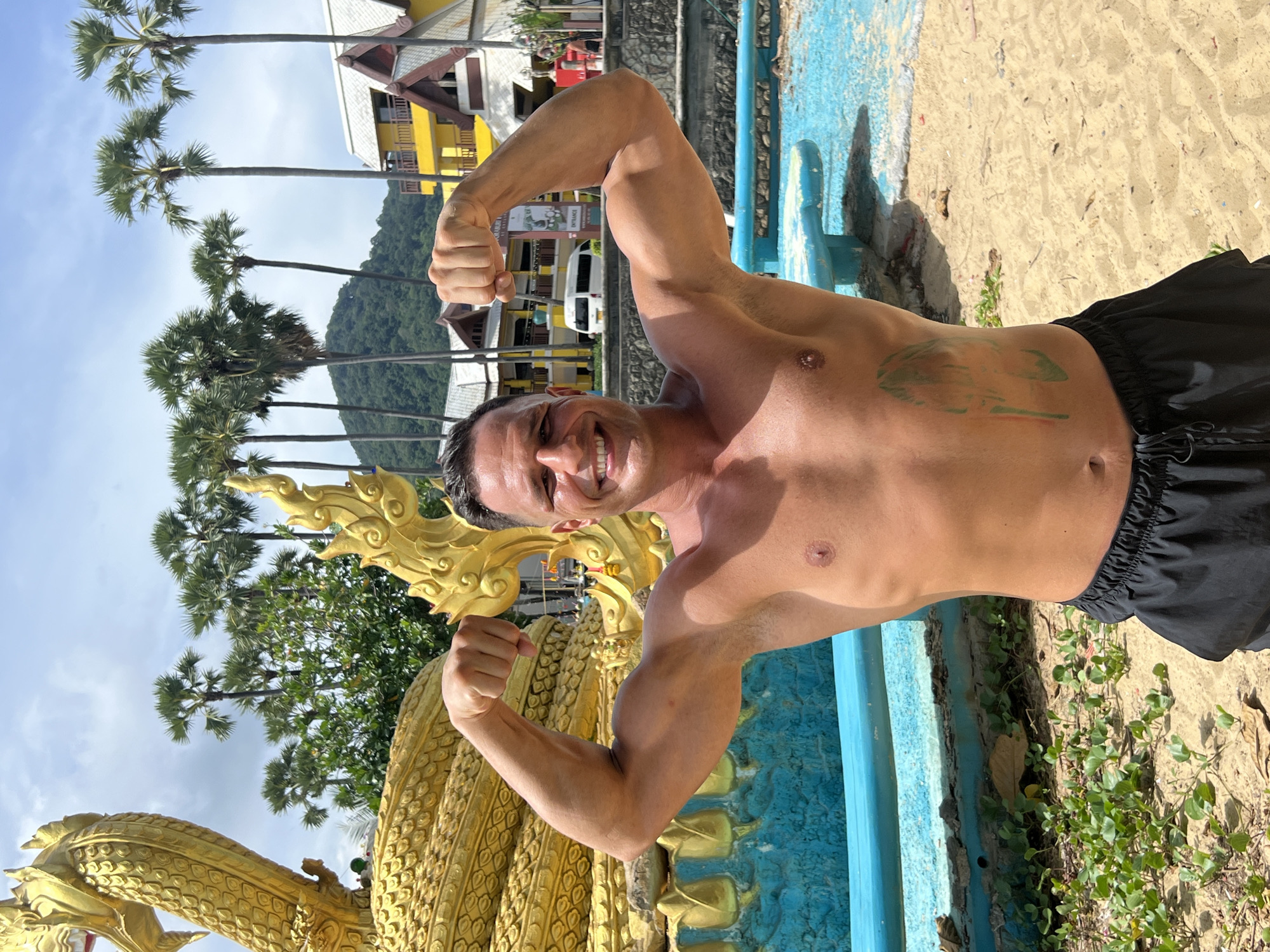 A shirtless man is flexing his biceps in front of a large, ornate golden dragon sculpture in Karon, Thailand. He is wearing black swim trunks. The sculpture is situated near a vibrant turquoise bench and a sandy beach area. In the background, several palm trees line the coast, with buildings and a green hillside visible further beyond. The sky is partly cloudy and the scene appears to be a sunny day.