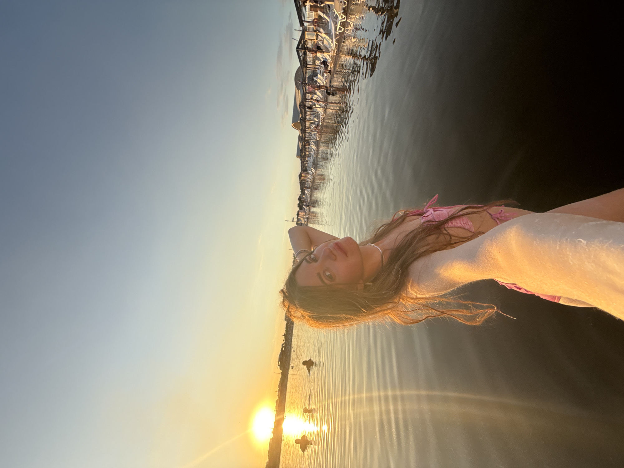 A young woman is taking a selfie in a body of water, likely a lake or salt lake, with the sunset in the background. She is wearing a pink bikini and has her arm raised, with her hand behind her head. The water reflects the golden light of the sunset. In the background, there are several people in the water and a shoreline with rows of lounge chairs and umbrellas, suggesting a beach resort or recreational area. The scene appears to be in Sol-Iletsk, Russia, known for its salt lakes and resorts. The sky transitions from a bright yellow near the horizon to a pale blue overhead.