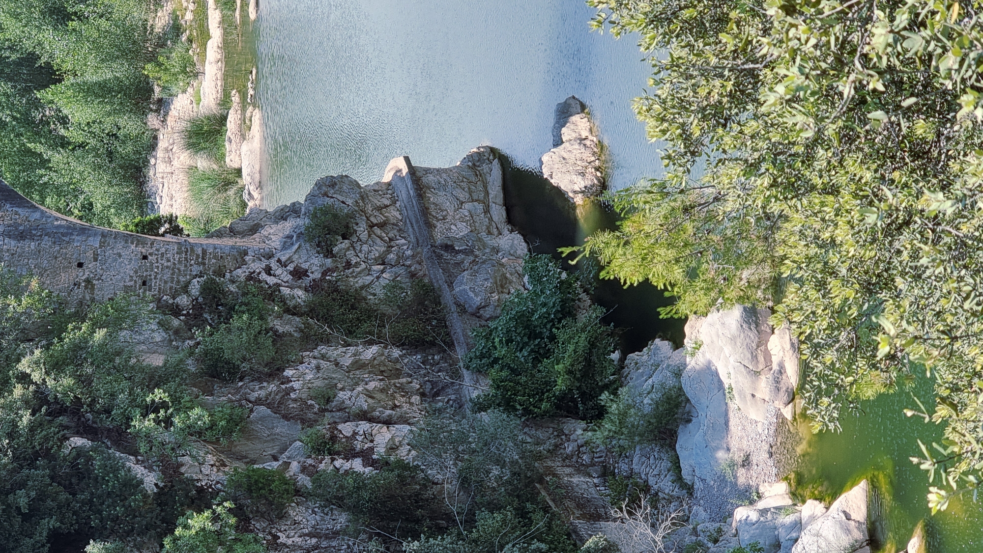 The image depicts a scenic view of the Pont del Liers, an ancient stone bridge spanning the river Muga in Tortellà, Spain. The bridge's weathered stone arches and masonry are visible, partially obscured by lush green vegetation and trees that cling to the rocky cliff face. A narrow pathway or road appears to follow the curve of the bridge, hugging the rock. The clear, calm waters of the river Muga are seen below, reflecting the surrounding landscape and the bright sky. The scene is bathed in natural sunlight, creating a serene and picturesque atmosphere.
